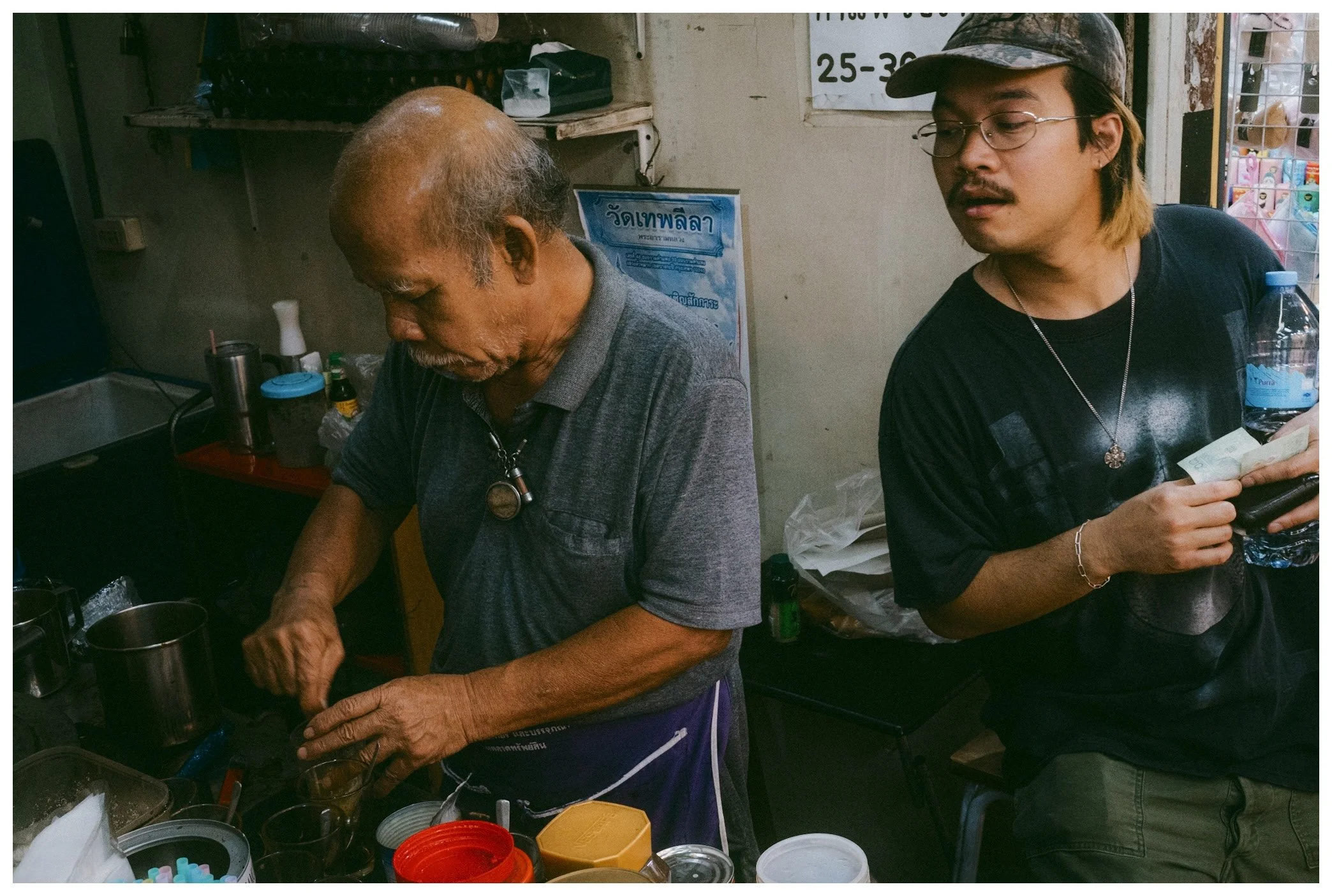 An elderly man and a younger man standing at a counter in a shop, with various small containers and bottles around them, possibly preparing or selling goods.