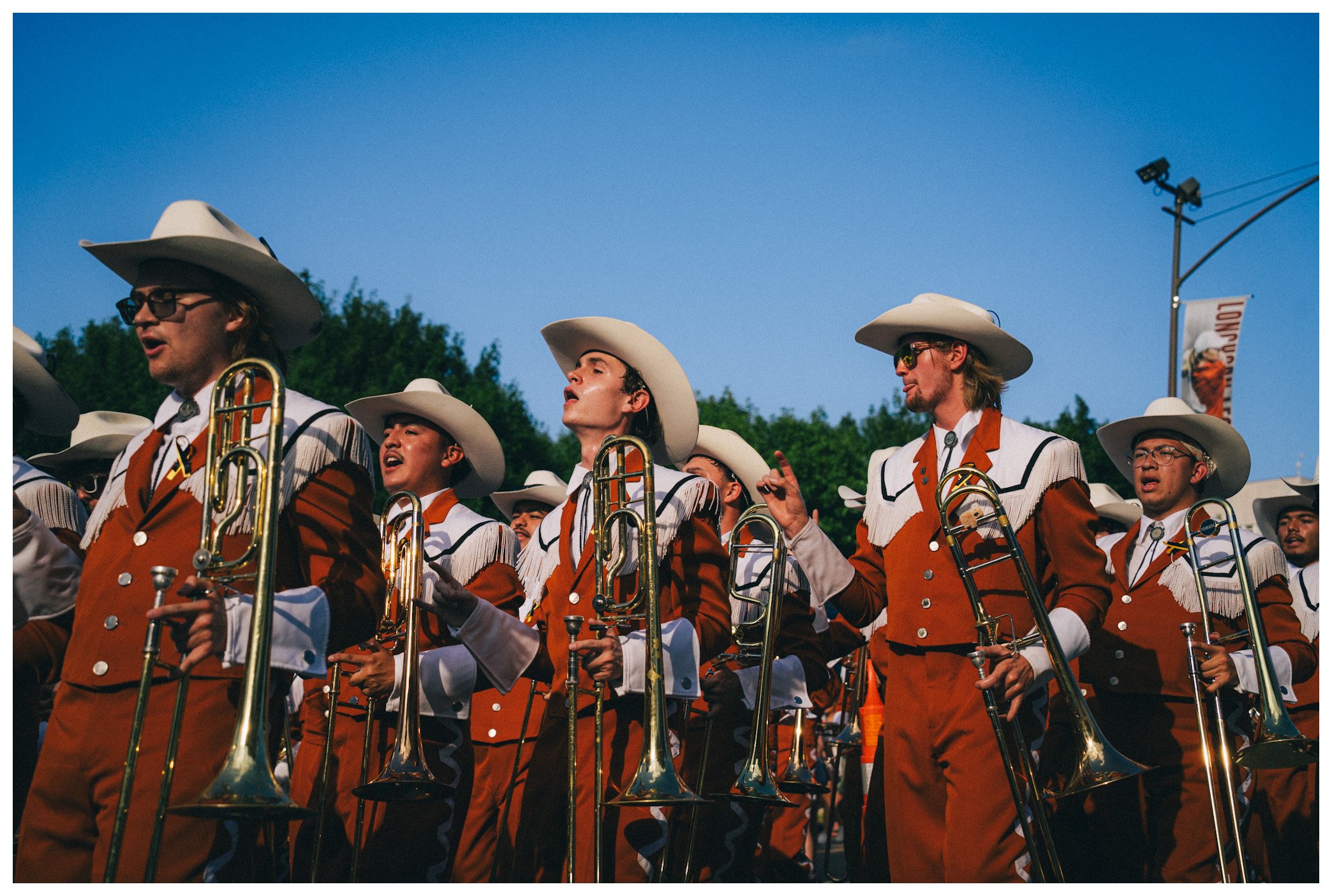 A marching band performing outdoors under a clear blue sky, with musicians wearing matching orange and white uniforms and white cowboy hats, playing brass instruments.