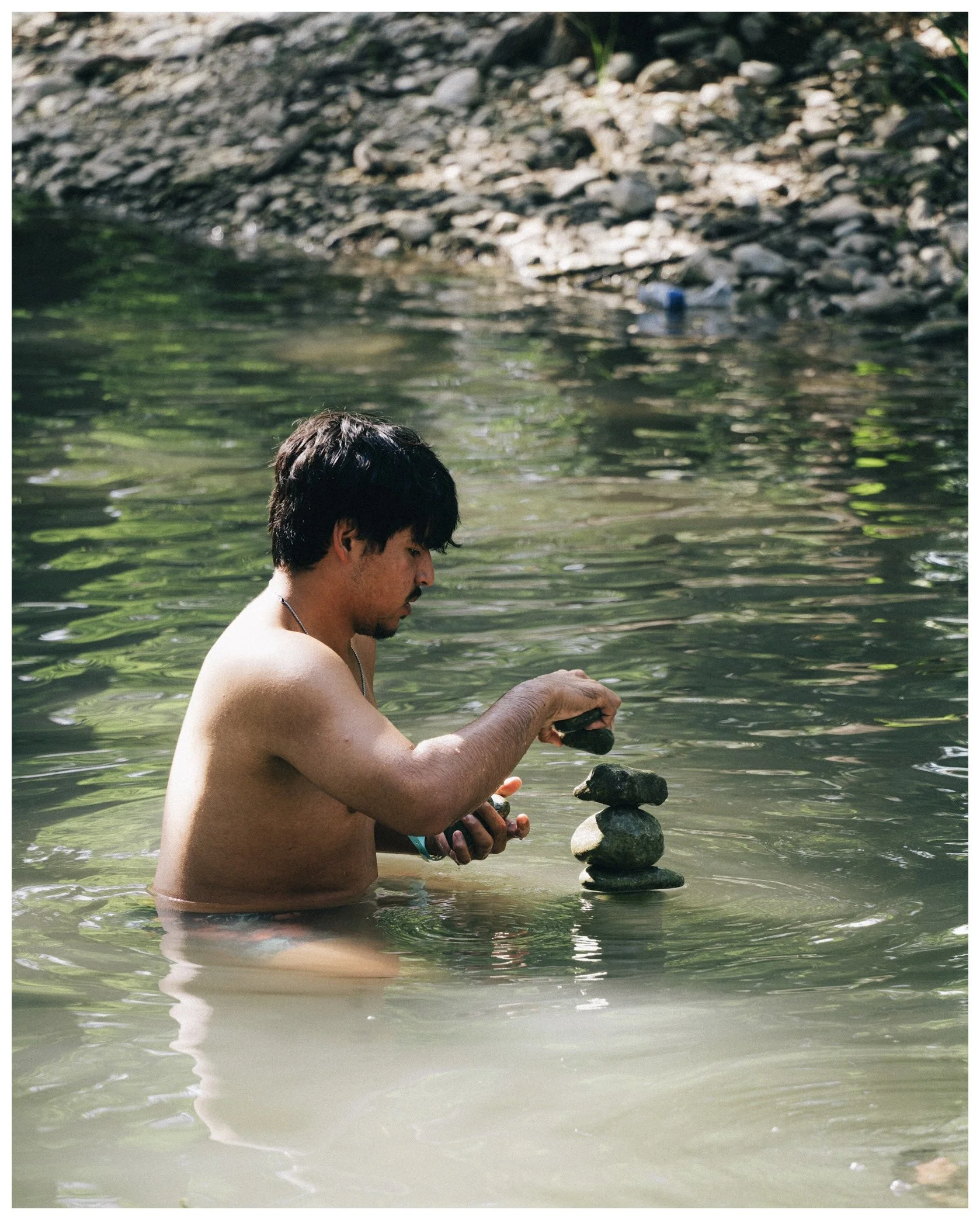 A shirtless young man with dark hair, standing waist-deep in a river, carefully stacking stones to create a balanced tower on top of the water.
