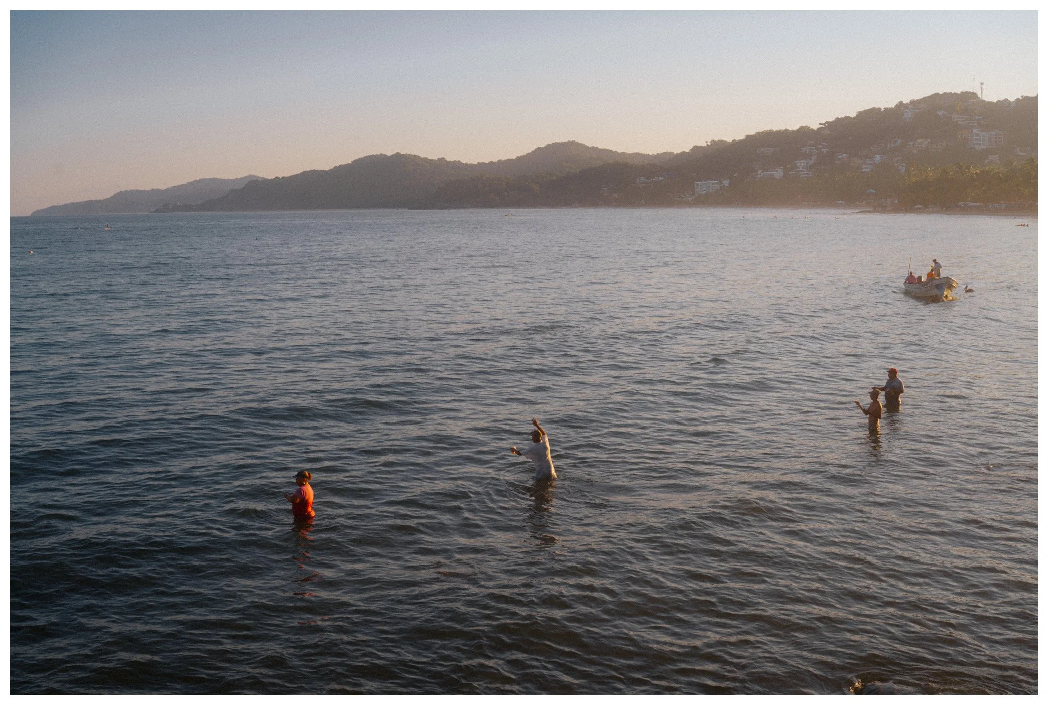 People fishing in the shallow waters near the shoreline with hills and houses in the background during sunset.