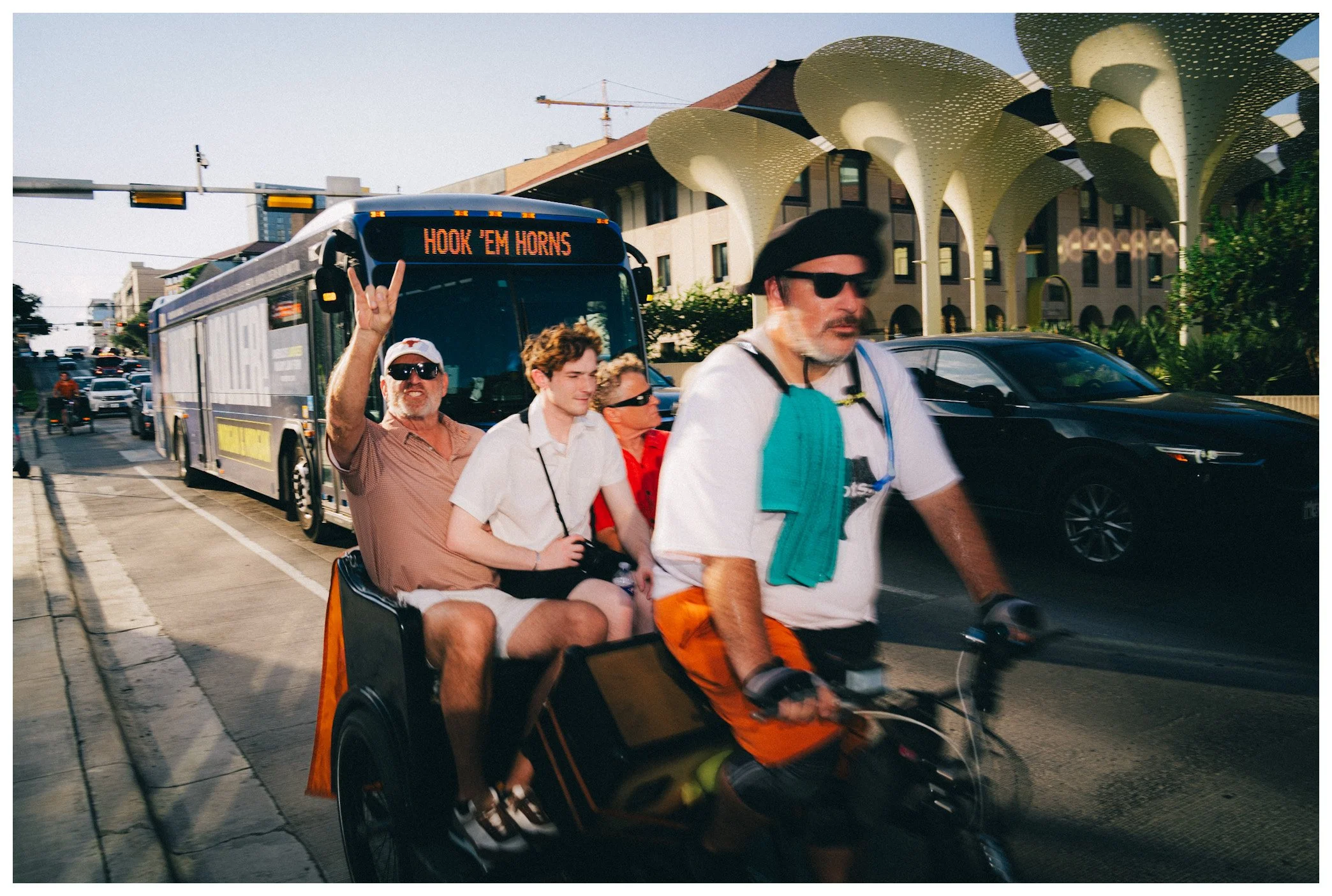 A group of four people riding a motorized bike or scooter in an urban area during daytime. One person is driving and three are seated behind. The person in the front is wearing black sunglasses, a white shirt, orange shorts, and a black helmet. The p