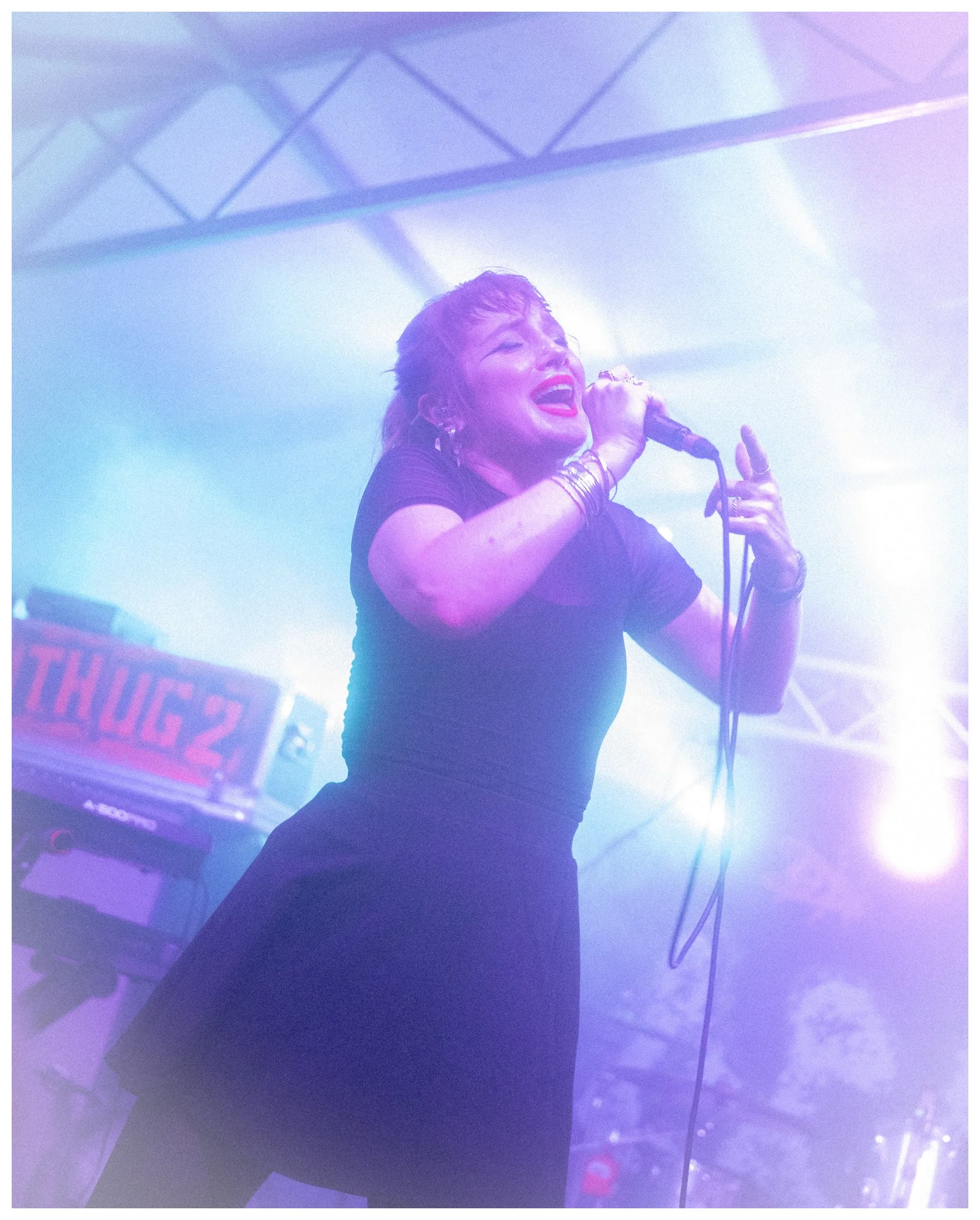 A woman singing into a microphone on stage with purple and blue lighting, wearing a black shirt, accessorized with rings and bracelets.