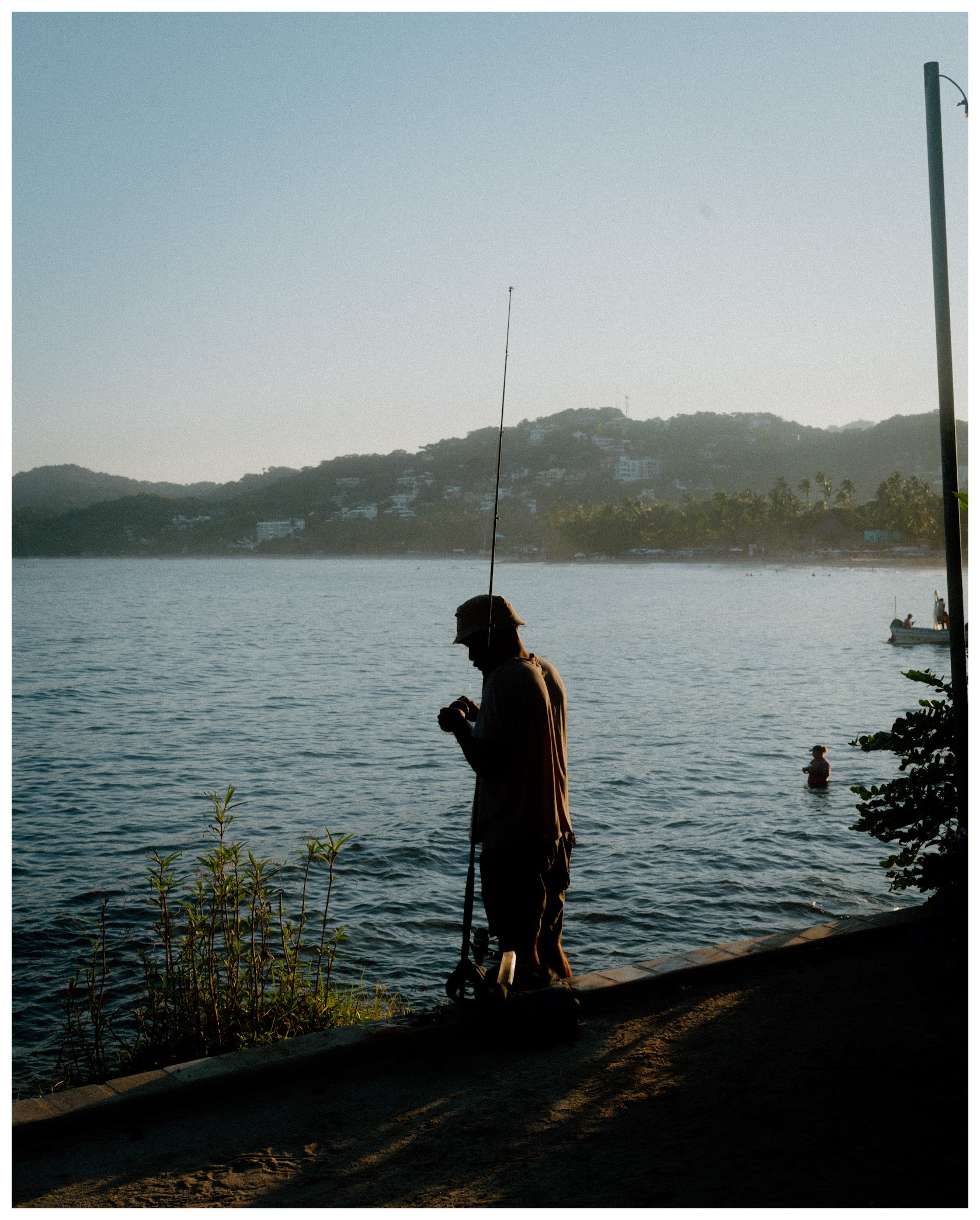 A person fishing by a body of water during sunset, with hills and houses in the background, and a boat with people fishing on the water.