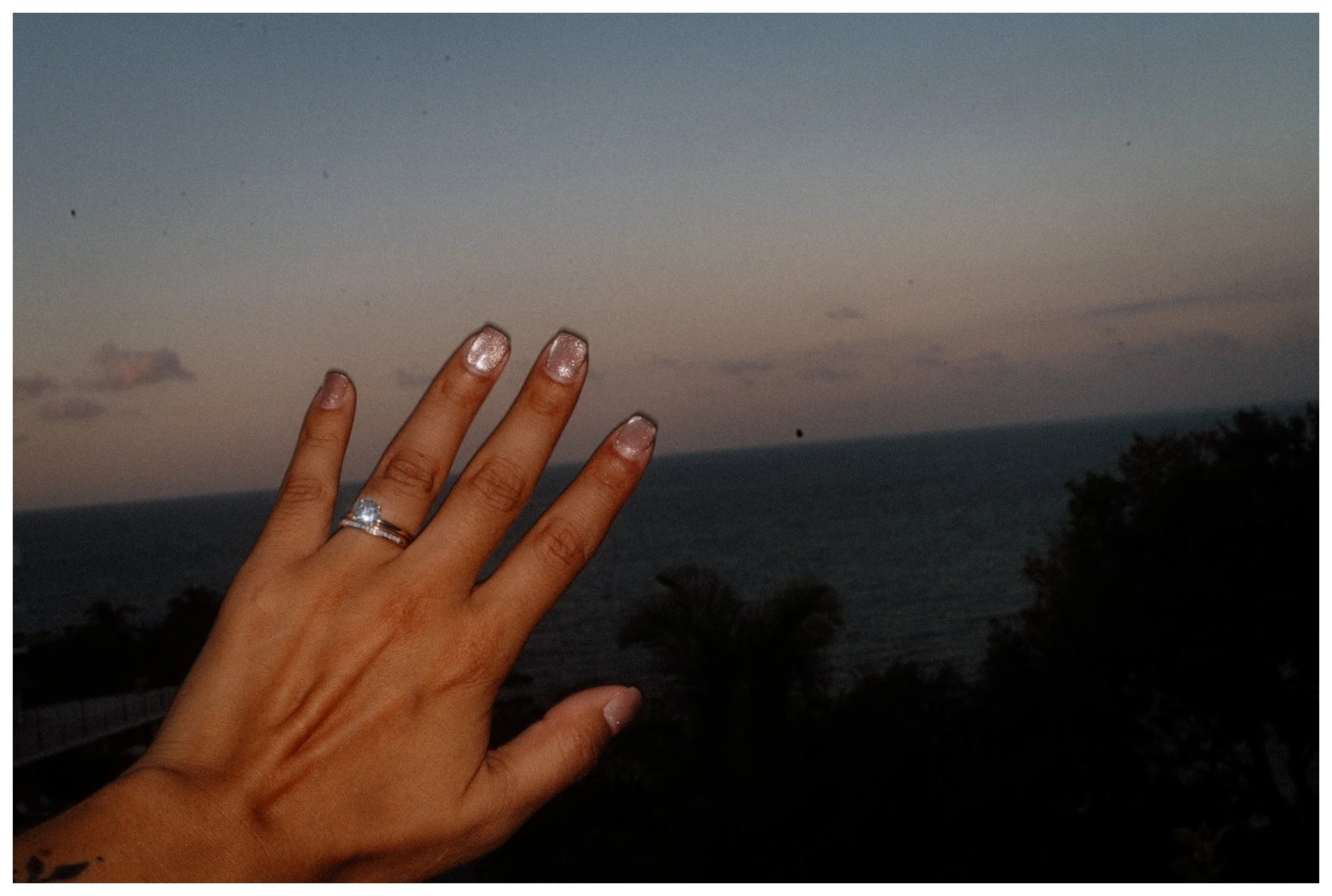 A person's left hand with a diamond engagement ring and a wedding band, held up against a sunset sky with clouds over a body of water and trees in the distance.
