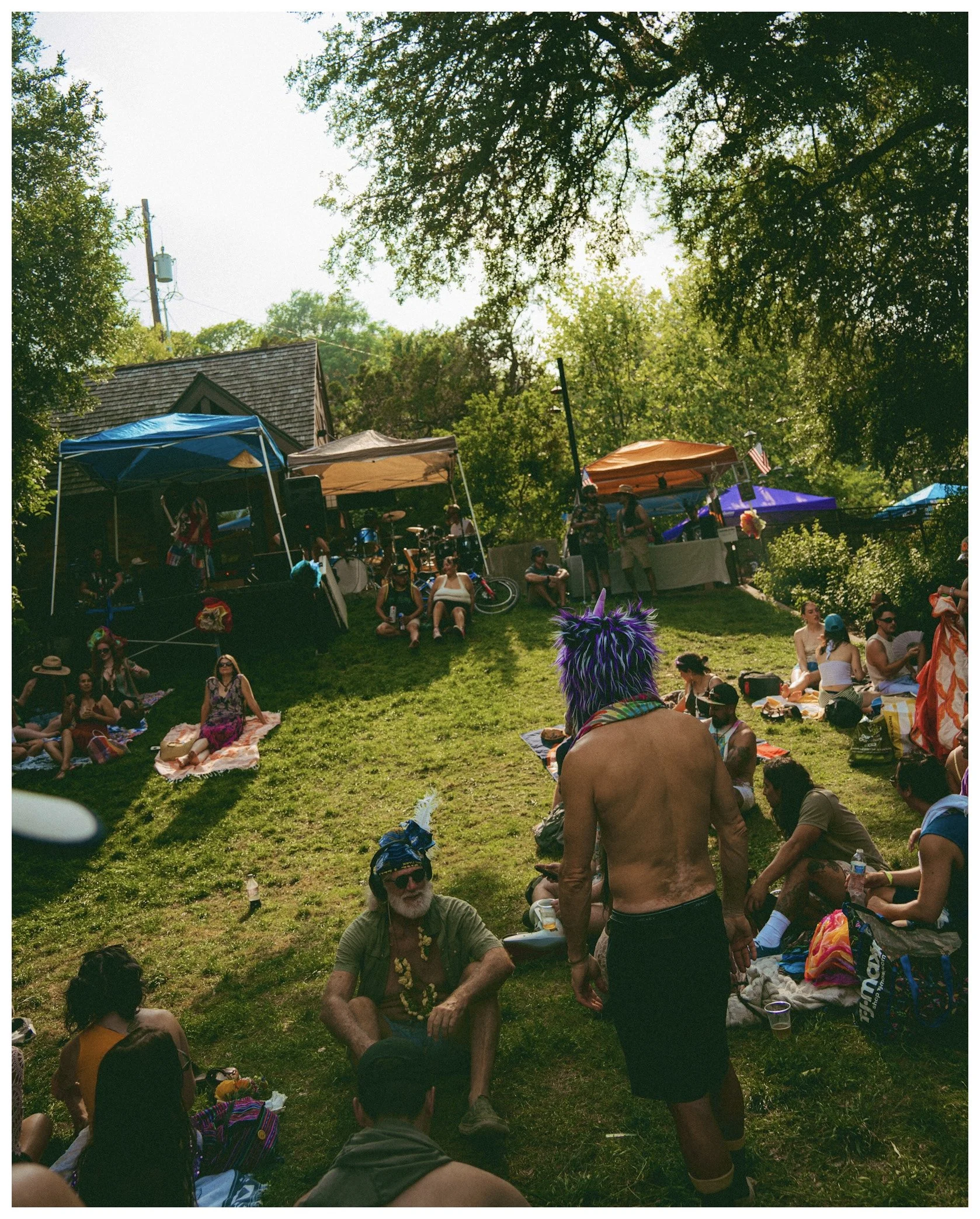 People gathered outdoors under trees, sitting and standing on a grassy slope, at a festival or outdoor event with tents and balloons.