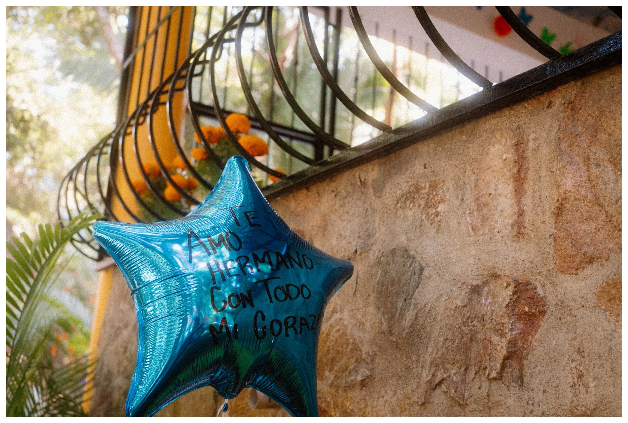 Blue star-shaped balloon with handwritten message in Spanish, hanging in front of a brick wall and black metal railing, with blurred orange flowers in the background.