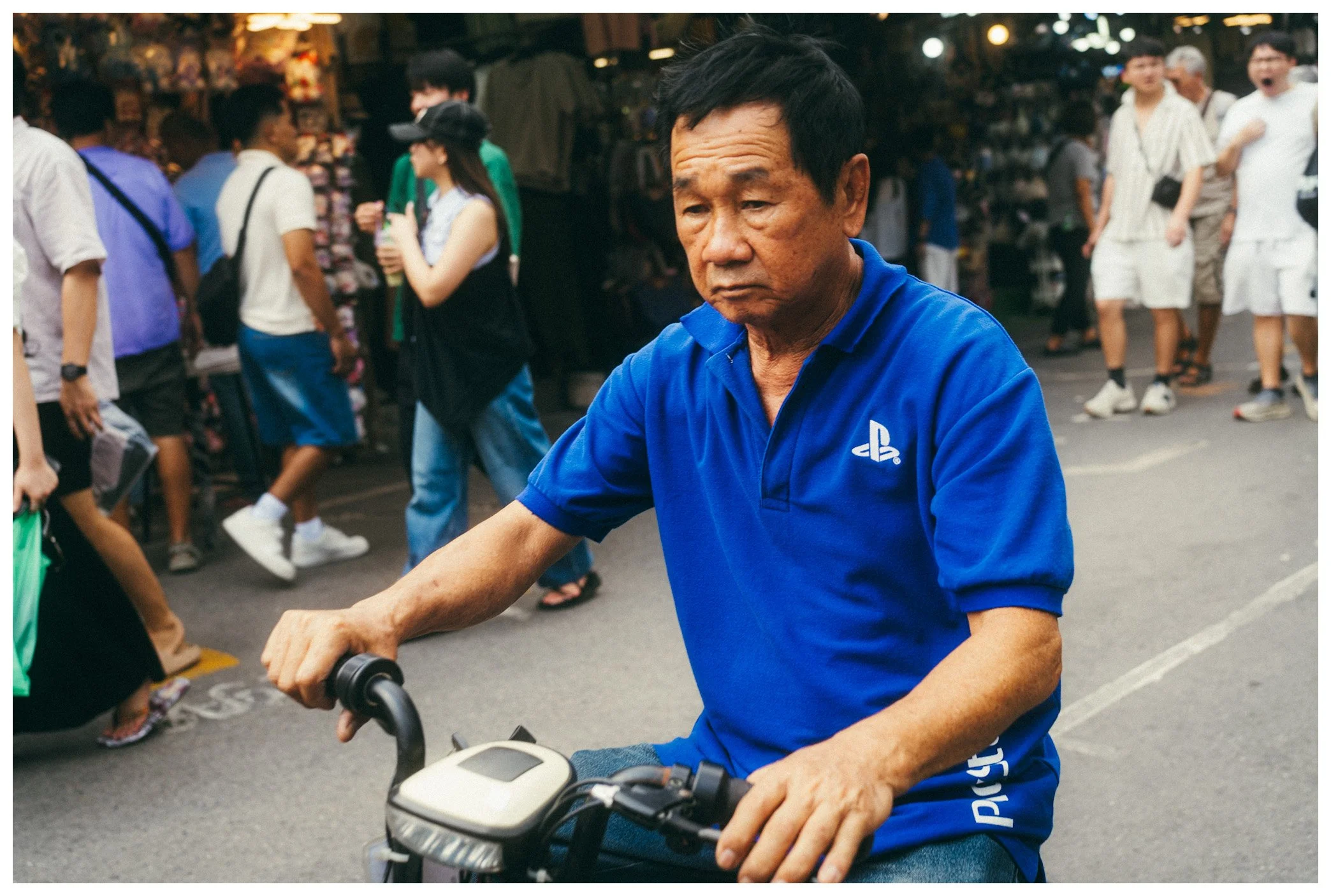 An elderly man in a blue polo shirt rides a bicycle in a busy outdoor market.