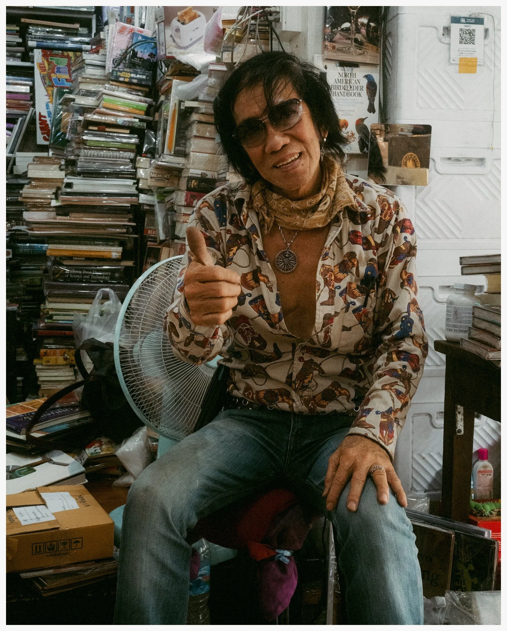 A man with dark hair wearing sunglasses, a patterned shirt, and a necklace, smiling and giving a thumbs up. He is sitting in a cluttered room filled with stacks of books, boxes, and various miscellaneous items.