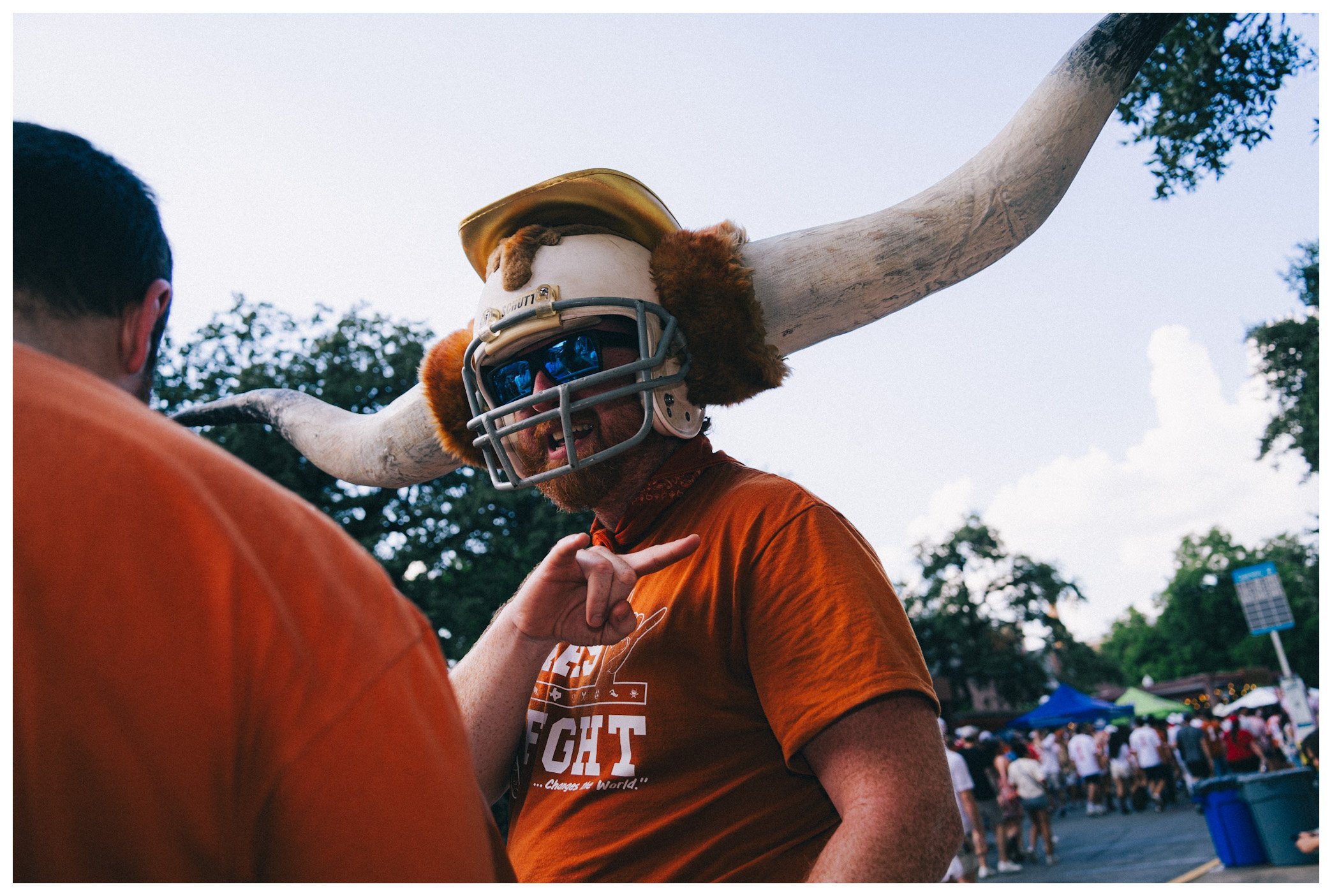 A man wearing sunglasses, a helmet, and an orange shirt, holding a large animal antler in a setting with trees and a crowd in the background.