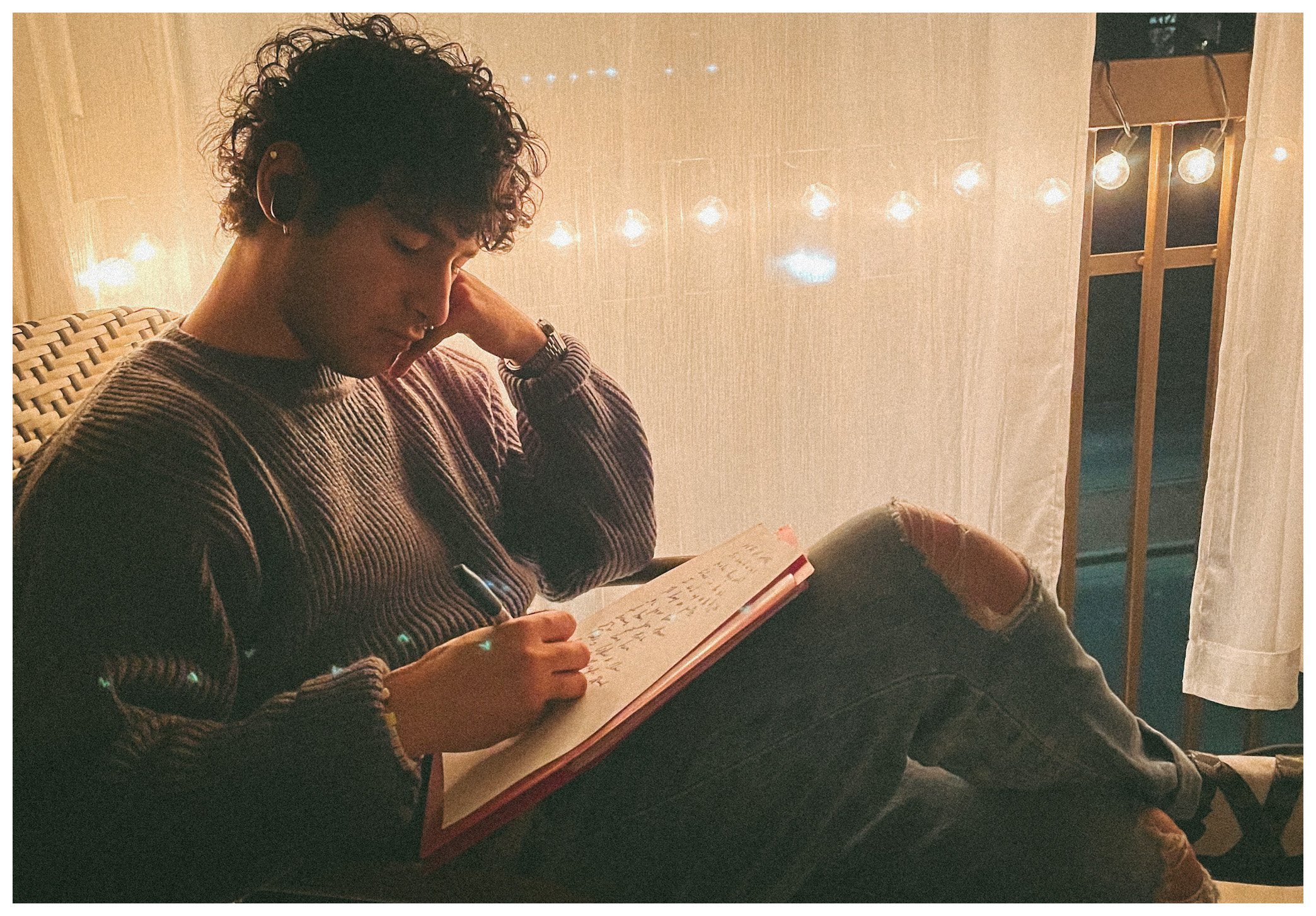 A young person with curly hair sitting on a chair, reading a book at night, with string lights and curtains in the background.