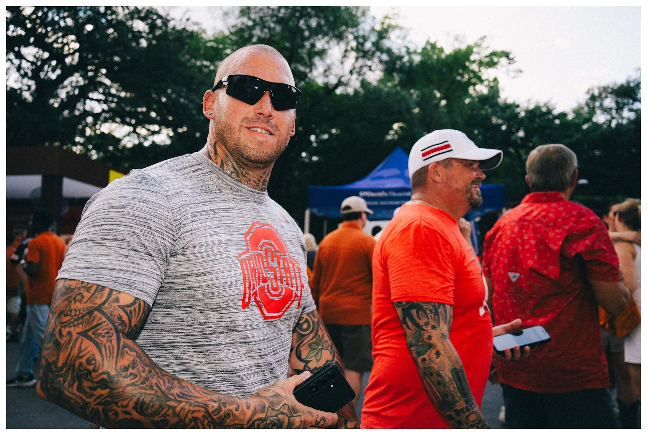 A man with tattoos wearing sunglasses and a gray Ohio State t-shirt standing outdoors among a crowd
