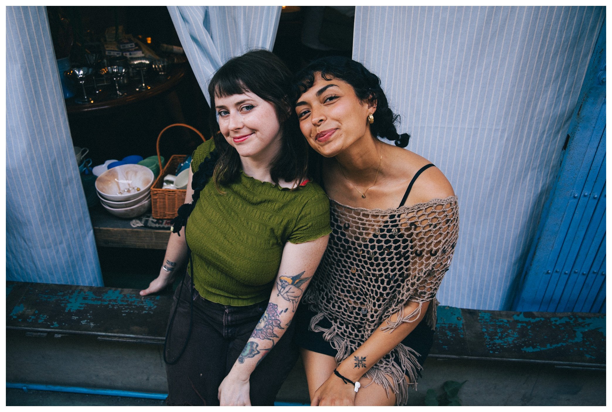 Two young women sitting closely together in a casual setting, smiling at the camera, with a background of shelves and blue-striped curtains.