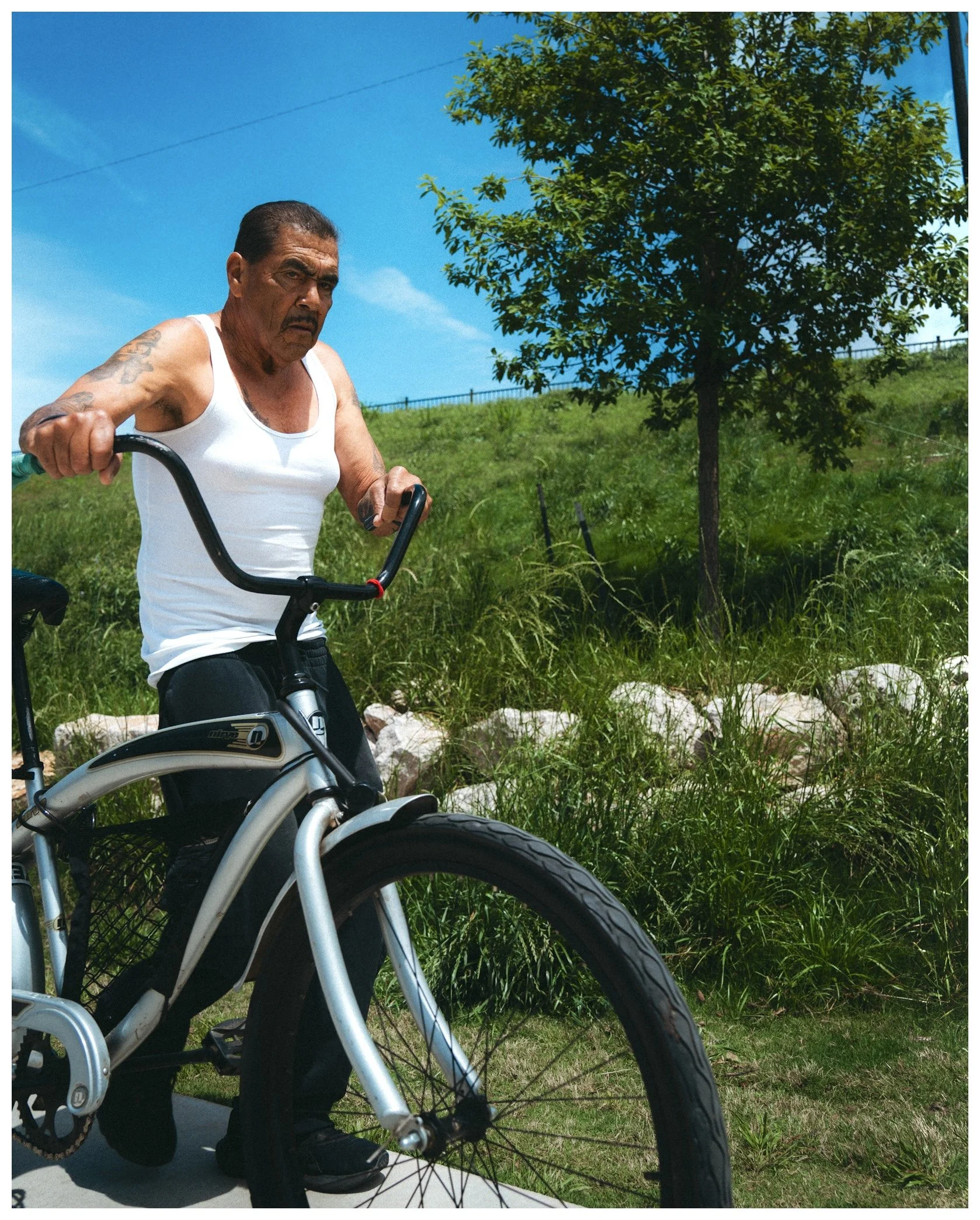 An elderly man with tattoos on his arm rides a motorized scooter outdoors on a sunny day, with green grass, a tree, and a bright blue sky in the background.
