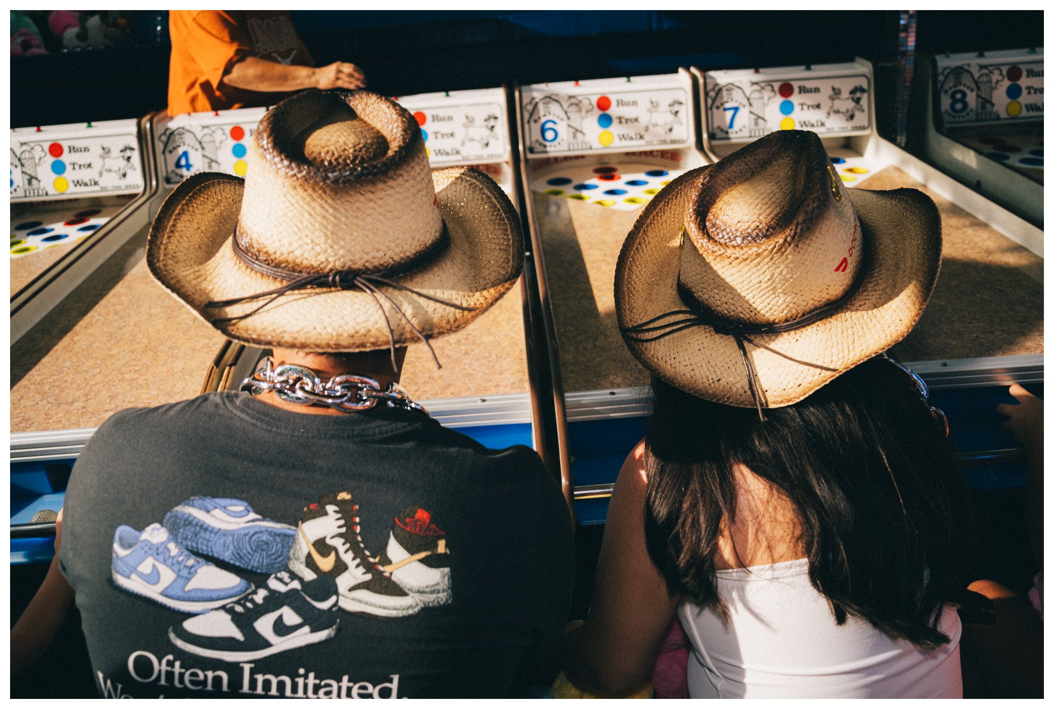 Two people wearing cowboy hats sitting at a carnival game with numbered sections and colored dots, with other people and game booths visible in the background.