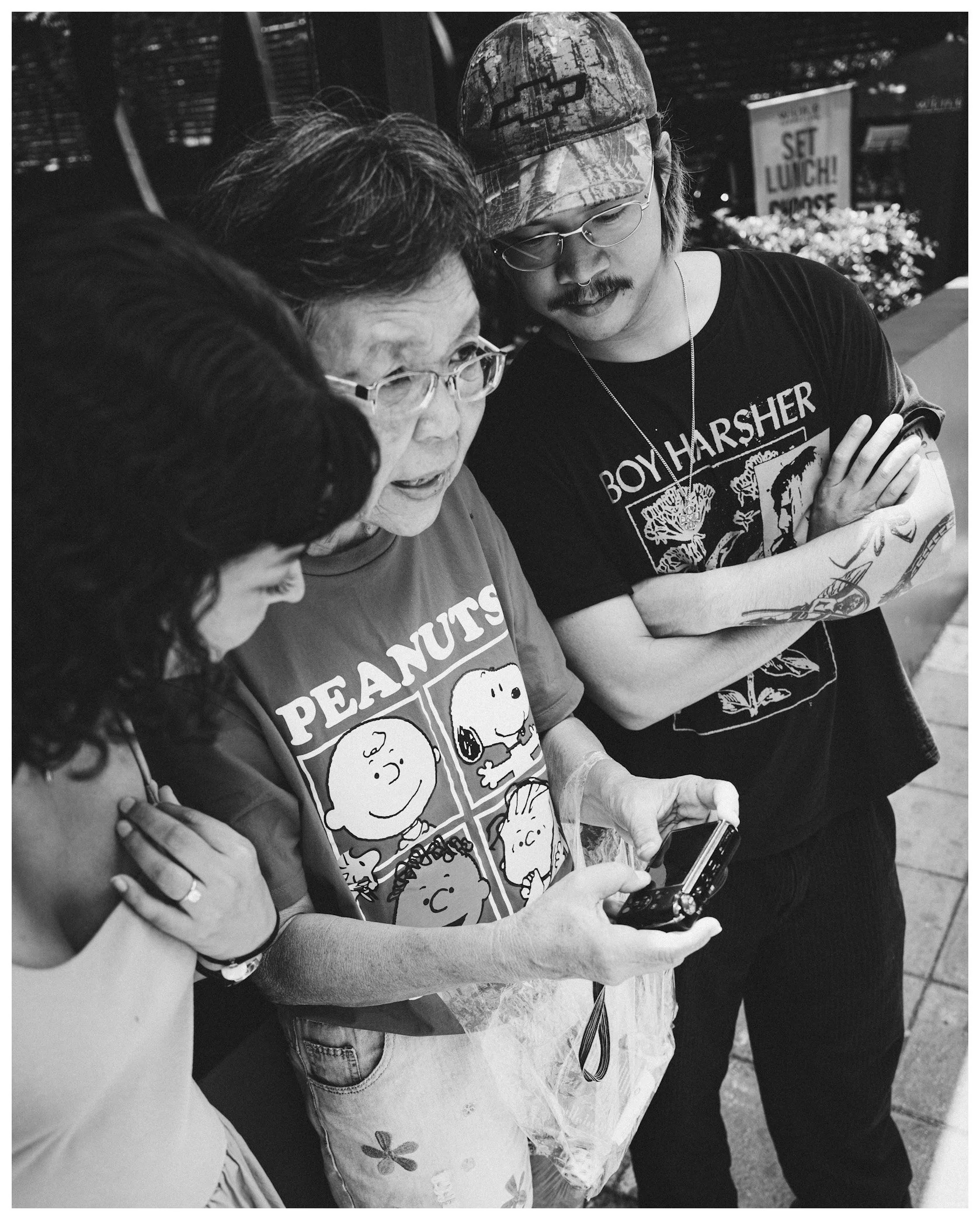 Four people huddled together looking at a smartphone, with a sign and outdoor decor in the background.