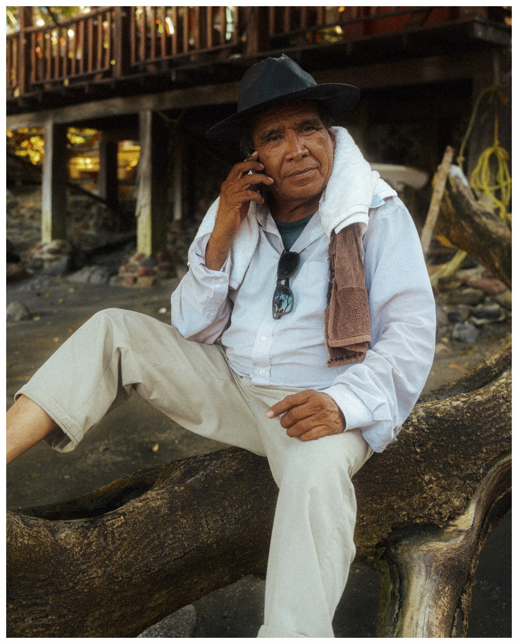 A man with dark curly hair, wearing a black hat, white shirt, and beige pants, sitting on a large tree branch with a white towel around his neck, holding a phone to his ear, in an outdoor setting with a wooden structure and autumn leaves in the backg