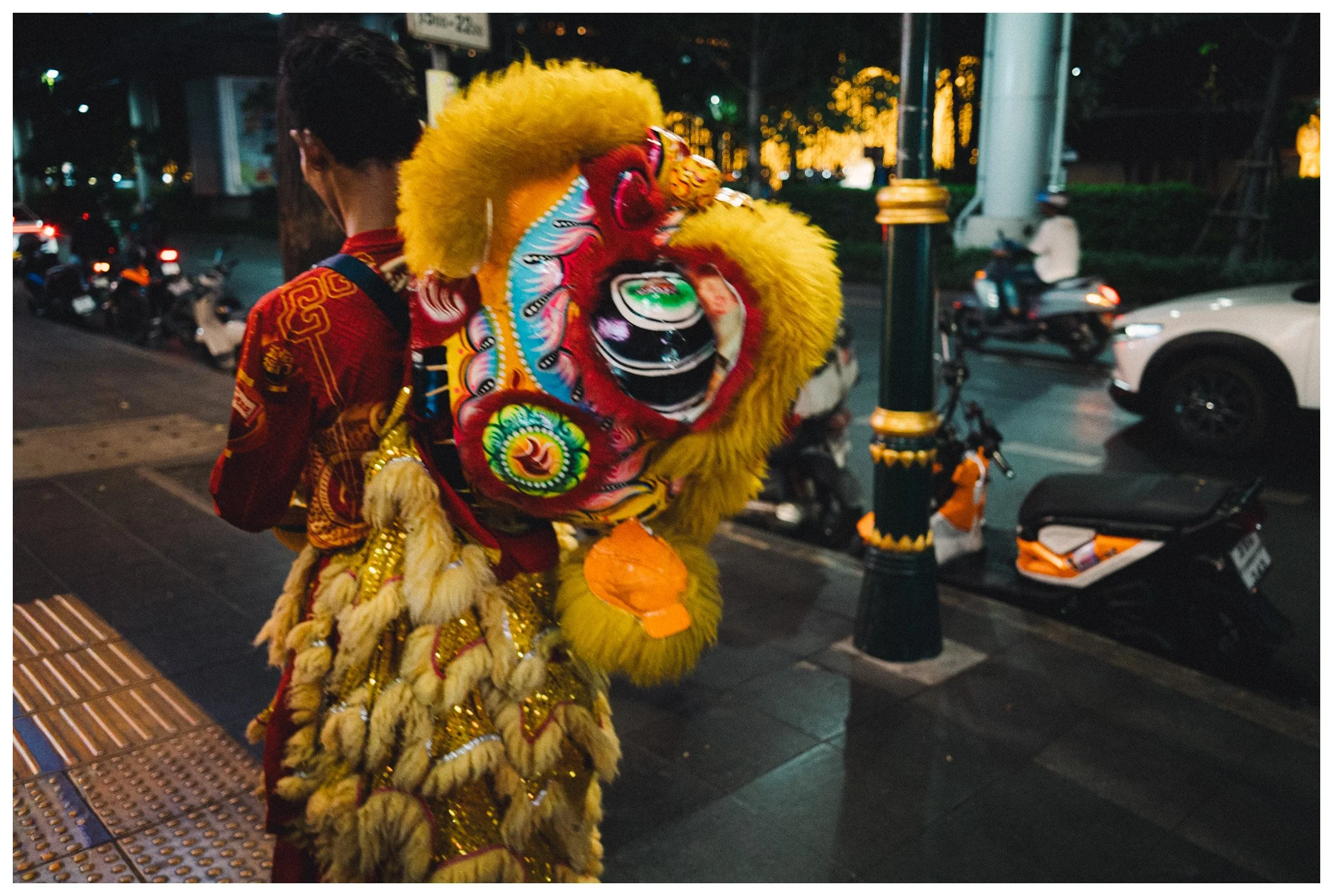 Person dressed in traditional Chinese lion dance costume standing on a city sidewalk at night, with parked scooters and cars in the background.