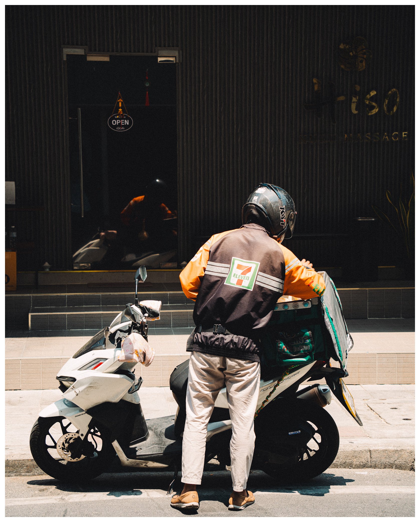 A person with a motorcycle helmet and beige pants standing next to a white scooter on a city street at night. A dark building with a sign that says "it's all" and a partially visible individual with a backpack are in the background.