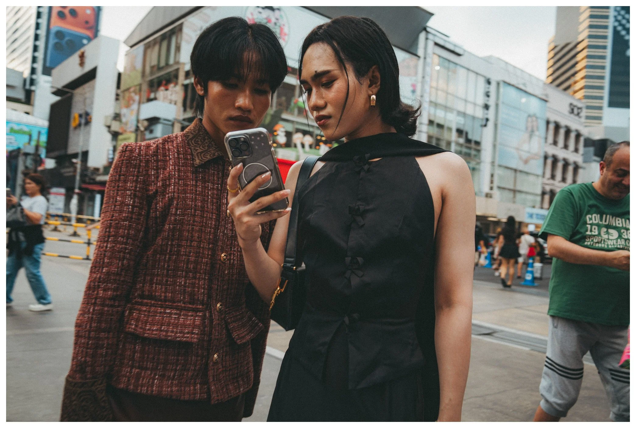 Two women are standing outside in a busy city, looking at a smartphone together. One woman has short black hair and is wearing a patterned blazer, while the other woman has shoulder-length black hair and is wearing a black sleeveless top. They seem f