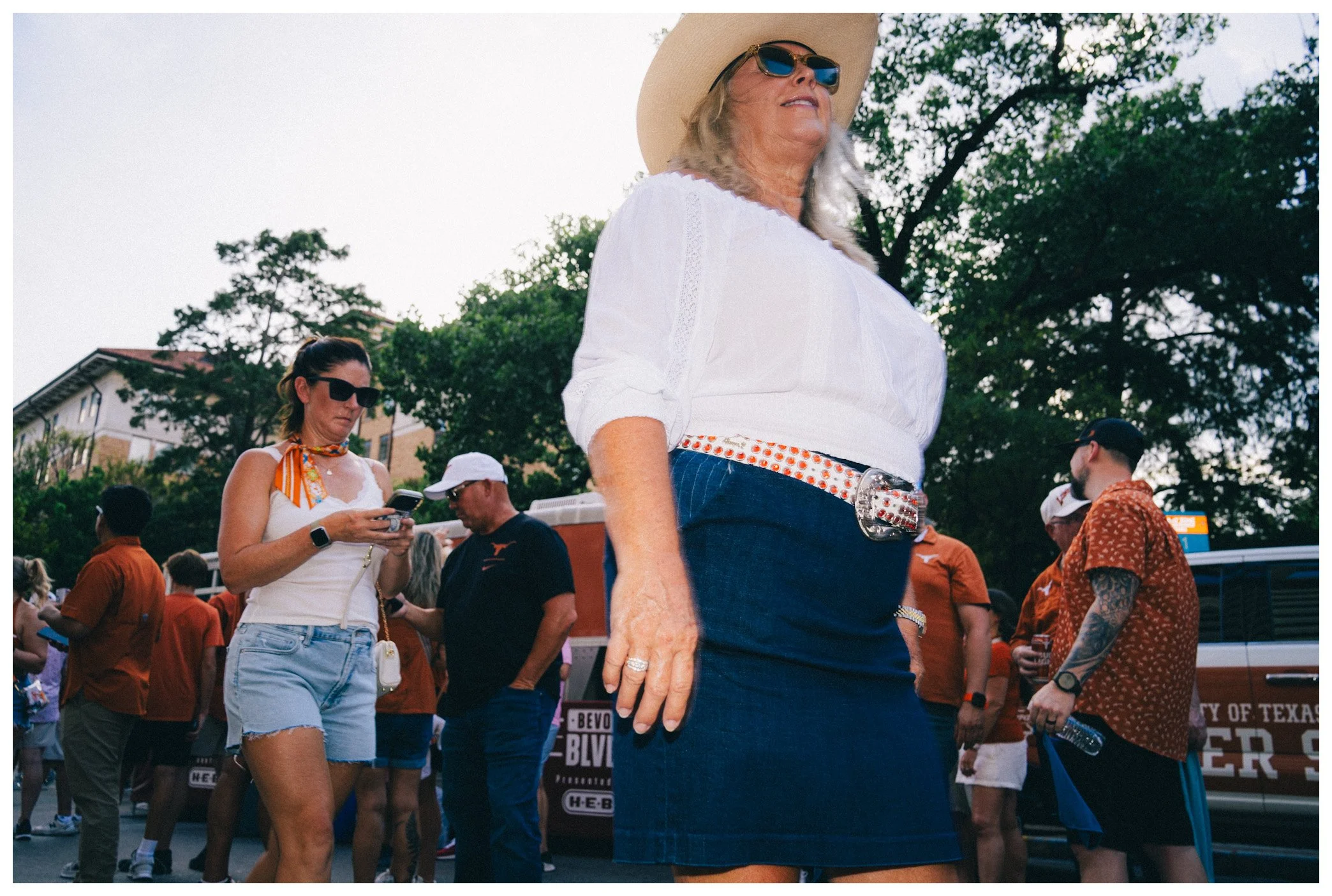 Close-up of a woman wearing a white blouse, denim skirt, cowboy hat, and sunglasses, at an outdoor event with several people and trees in the background.