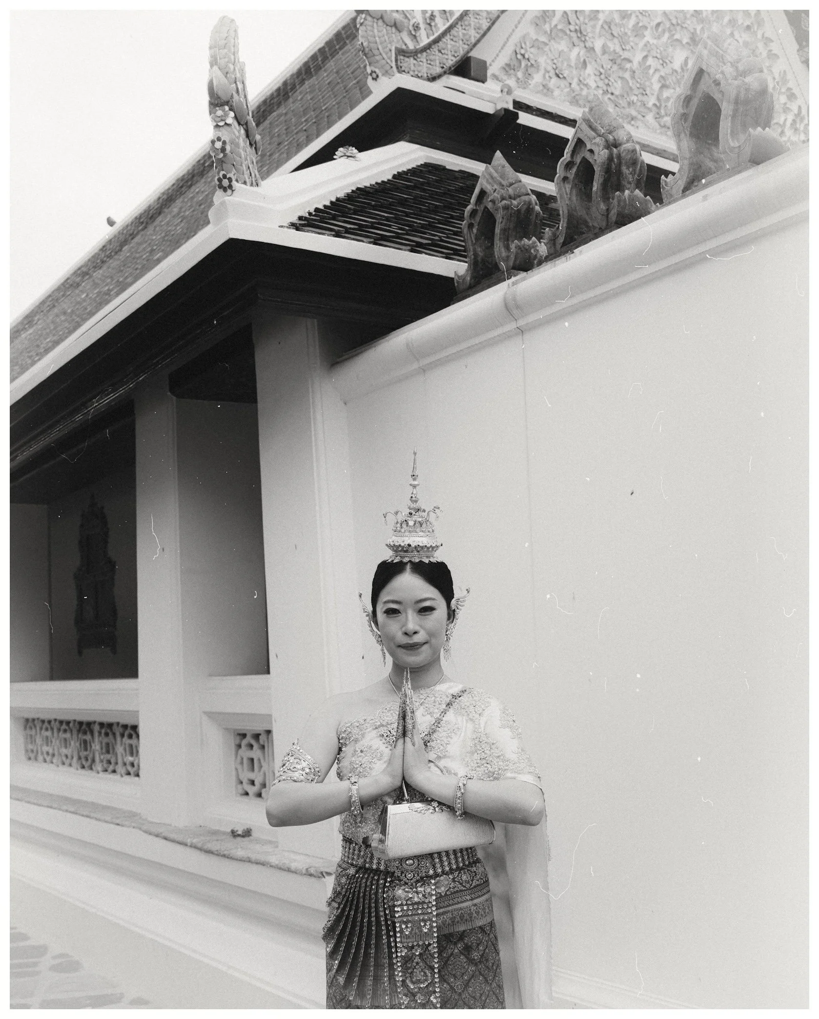 A woman dressed in traditional Thai attire standing indoors with her hands pressed together in a prayer pose. She is wearing a detailed, patterned Thai dress and a tall, ornate headdress. The background features traditional Thai architecture with dec