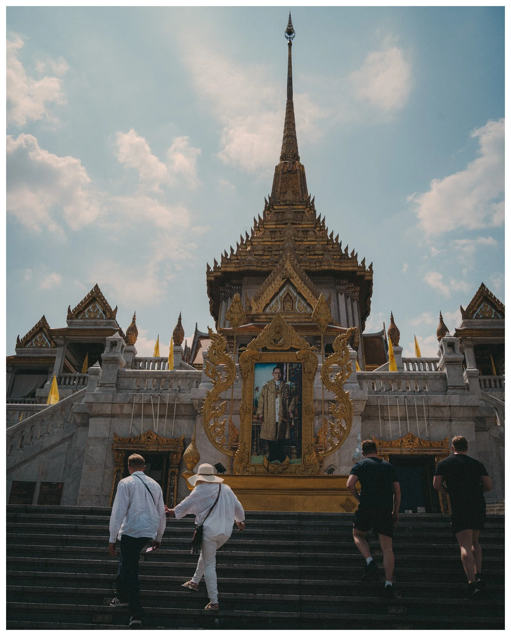 People walking up the stairs towards a decorated temple entrance with golden accents and a large portrait of a royal figure at the front, under a blue sky with clouds.