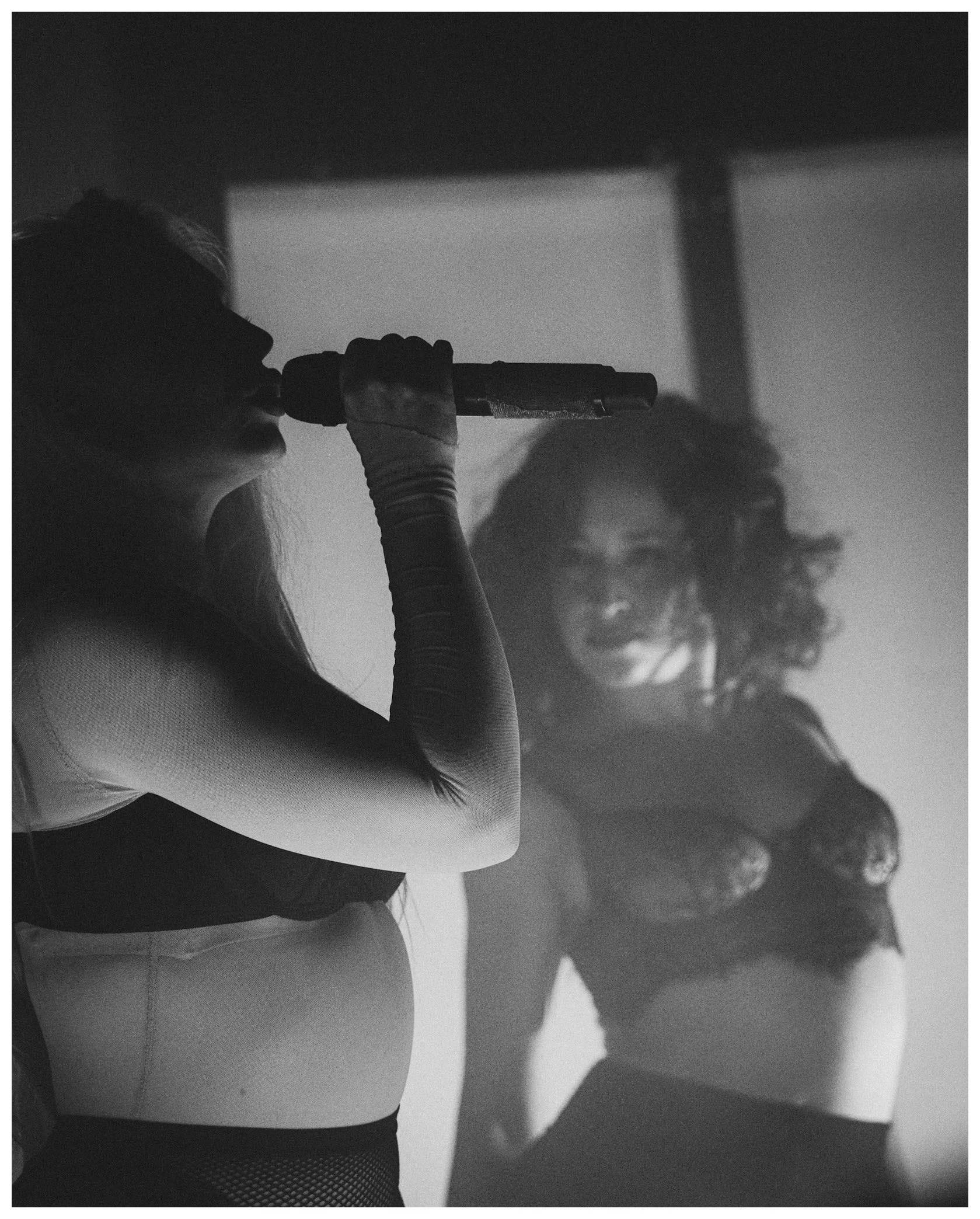 A person in athletic clothing and fishnet tights is drinking water from a bottle. The image is black and white, showing only the upper body and face, with curly hair partly visible.