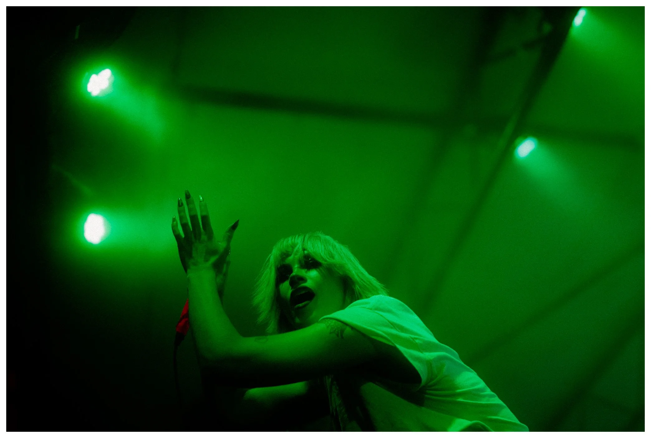 A woman with blonde hair and makeup is singing or shouting on stage, surrounded by green stage lighting, with her hand raised and mouth open.
