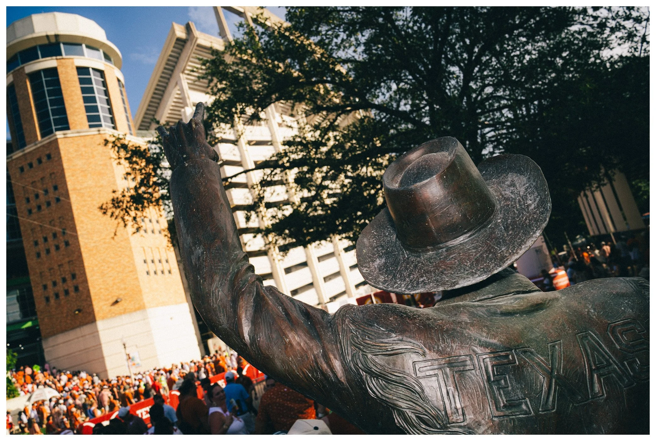 A bronze statue of a person with a top hat, raising their hand, is set against a cityscape with a crowd of people in the background, including high-rise buildings and trees.