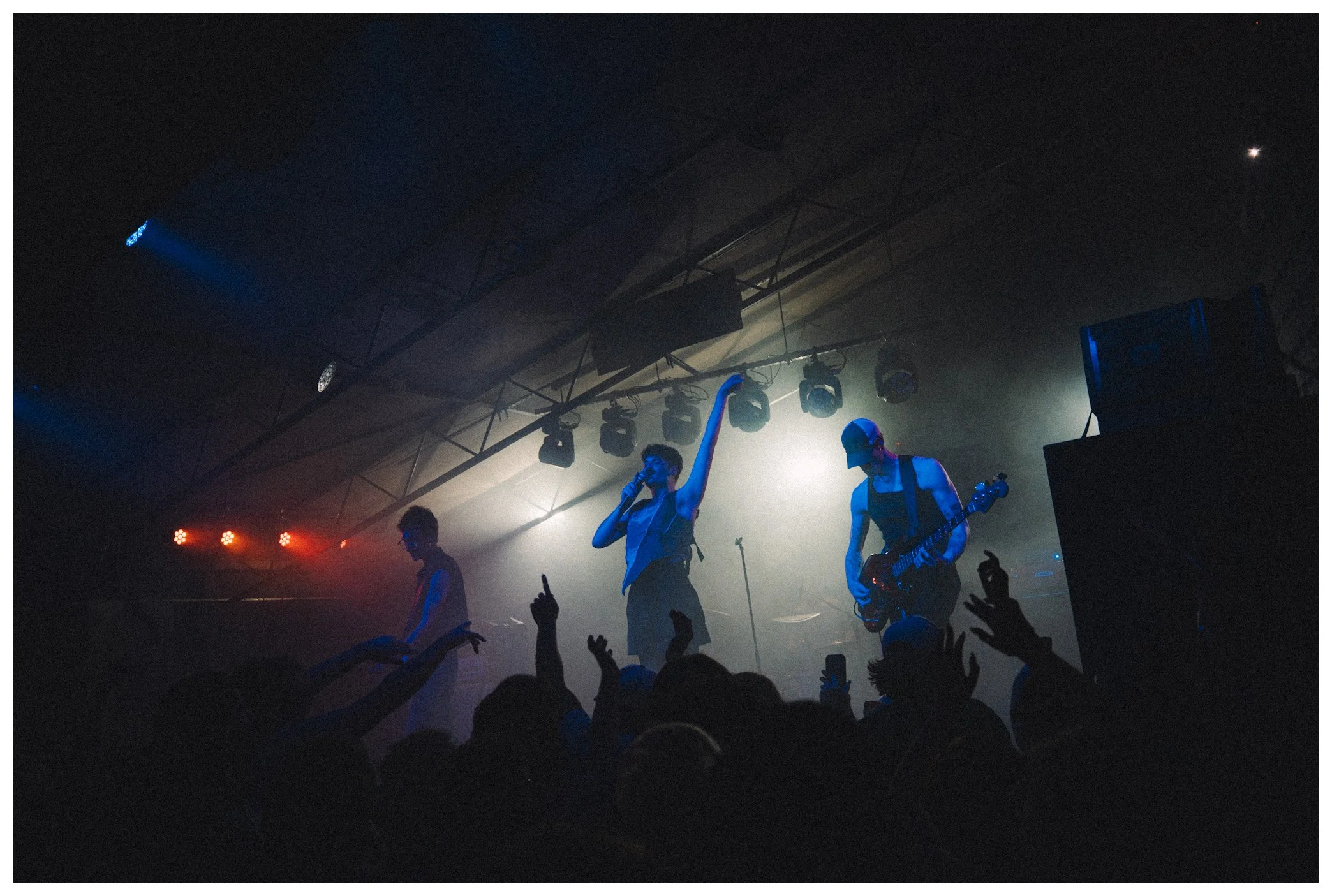 Musicians performing on stage at a concert with audience raising their hands. Stage lighting creates a moody atmosphere.