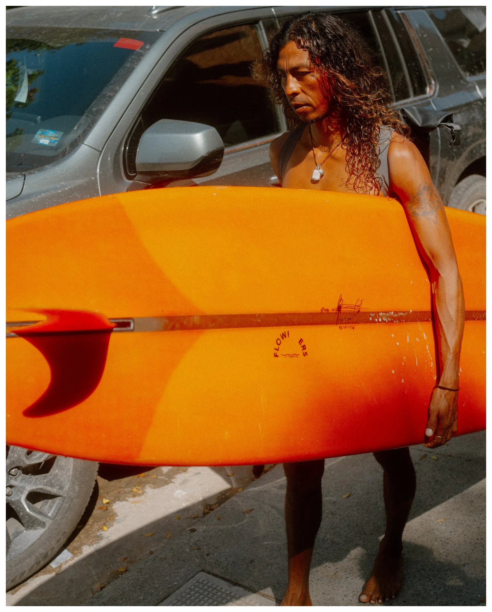 Person standing with a surfboard in front of a vehicle on a street.