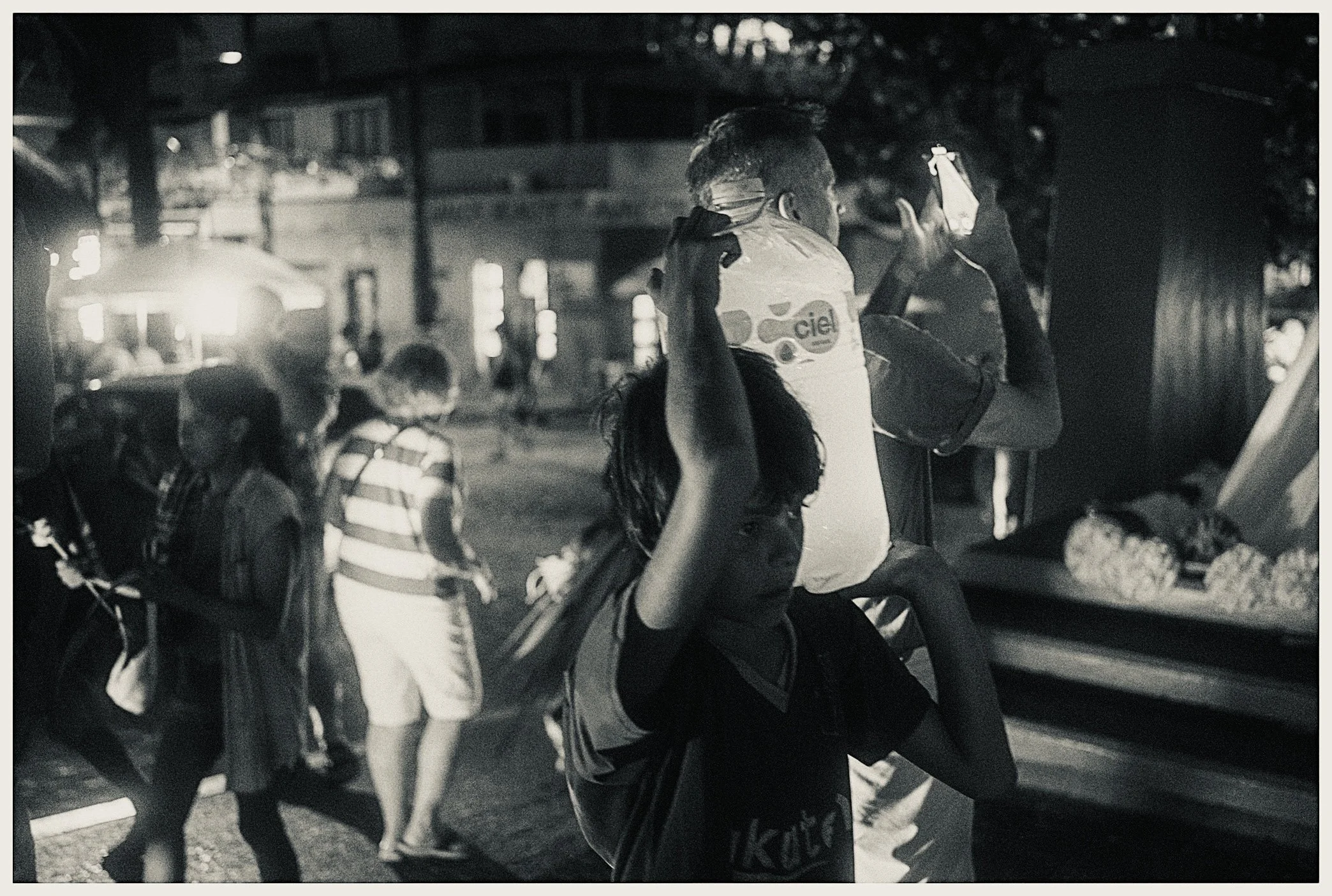 A woman with long hair wearing a striped shirt interacts with a person with short hair in a crowd at night, with several children and other people around, in an outdoor urban setting with buildings and streetlights.