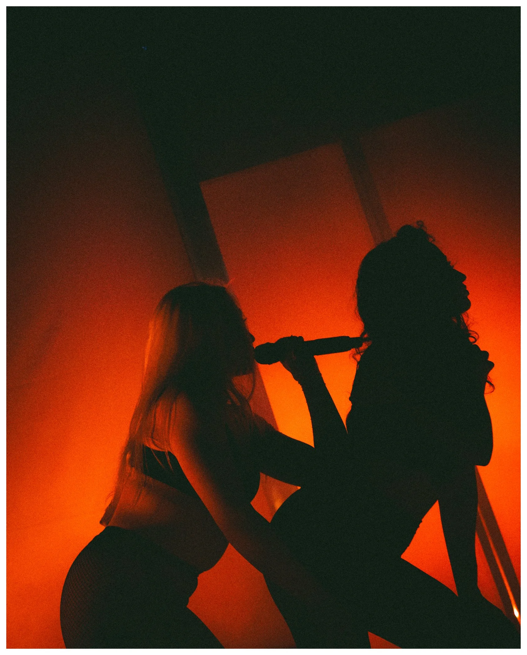 Silhouetted figures of a woman and a man against an orange background, engaging in an intimate moment with the woman leaning on the man's shoulder and the man resting his hand on her hip.