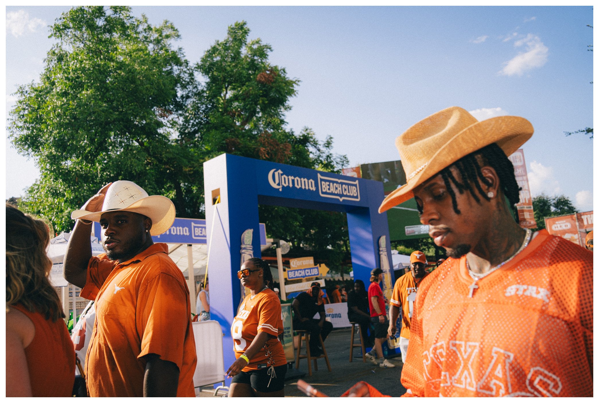 Young man with dreadlocks wearing an orange Texas Longhorns shirt and straw hat at a Corona Beach Club event, surrounded by other people in similar attire, under a clear blue sky with green trees in the background.