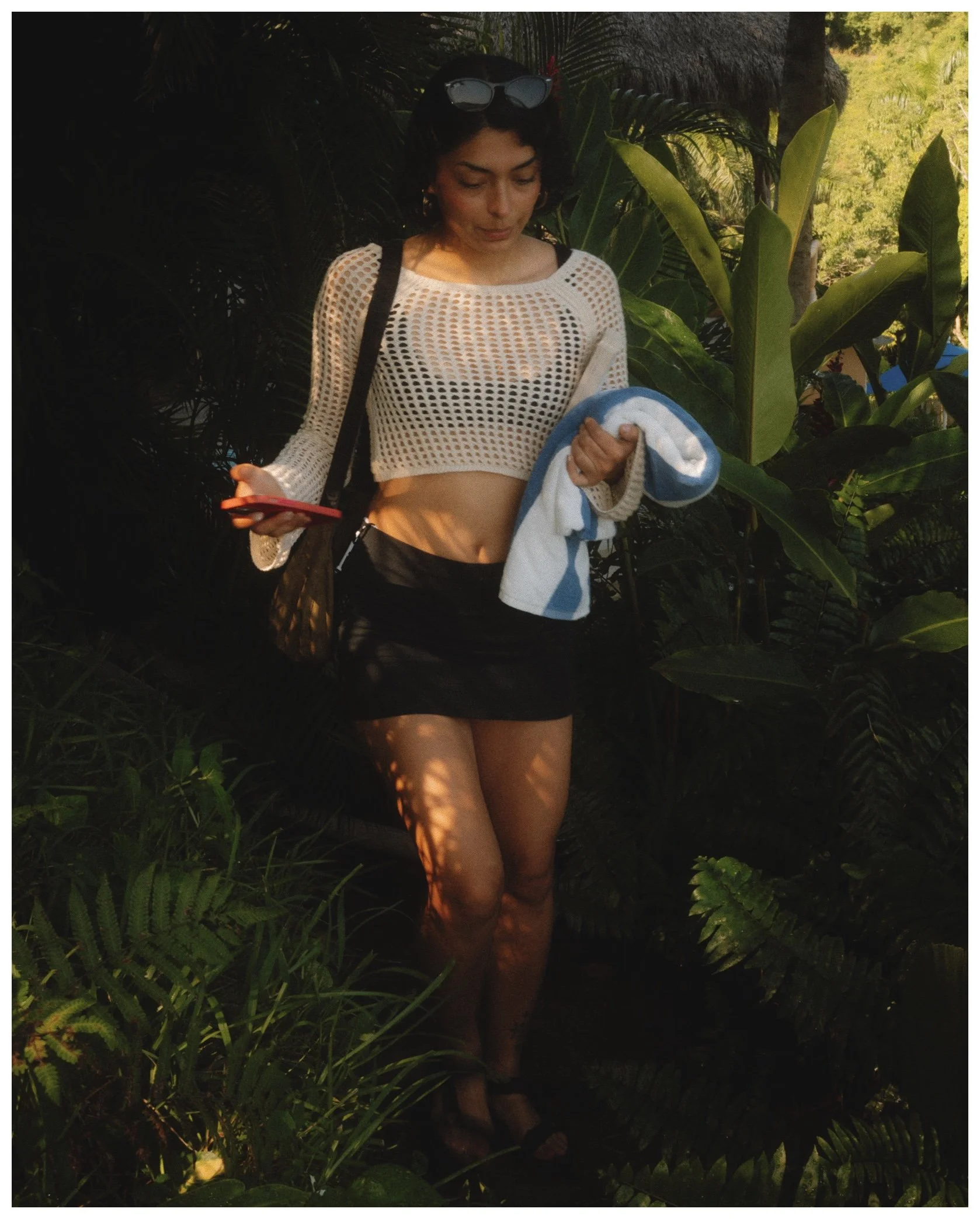 Woman standing among lush tropical plants, wearing a white crocheted crop top, black shorts, sunglasses on her head, and holding a towel.