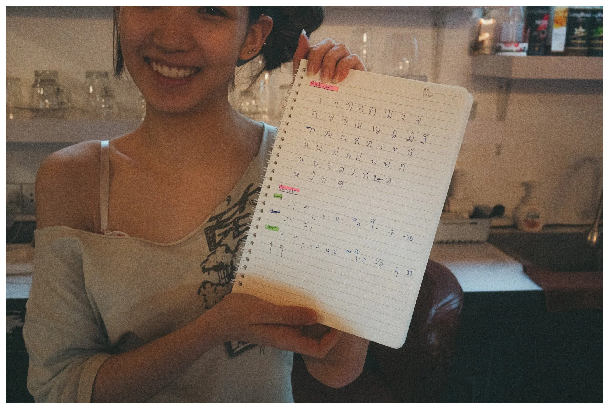 A person smiling and holding a notebook with Thai script, showing a portion of the alphabet, in a room with shelves of glassware in the background.