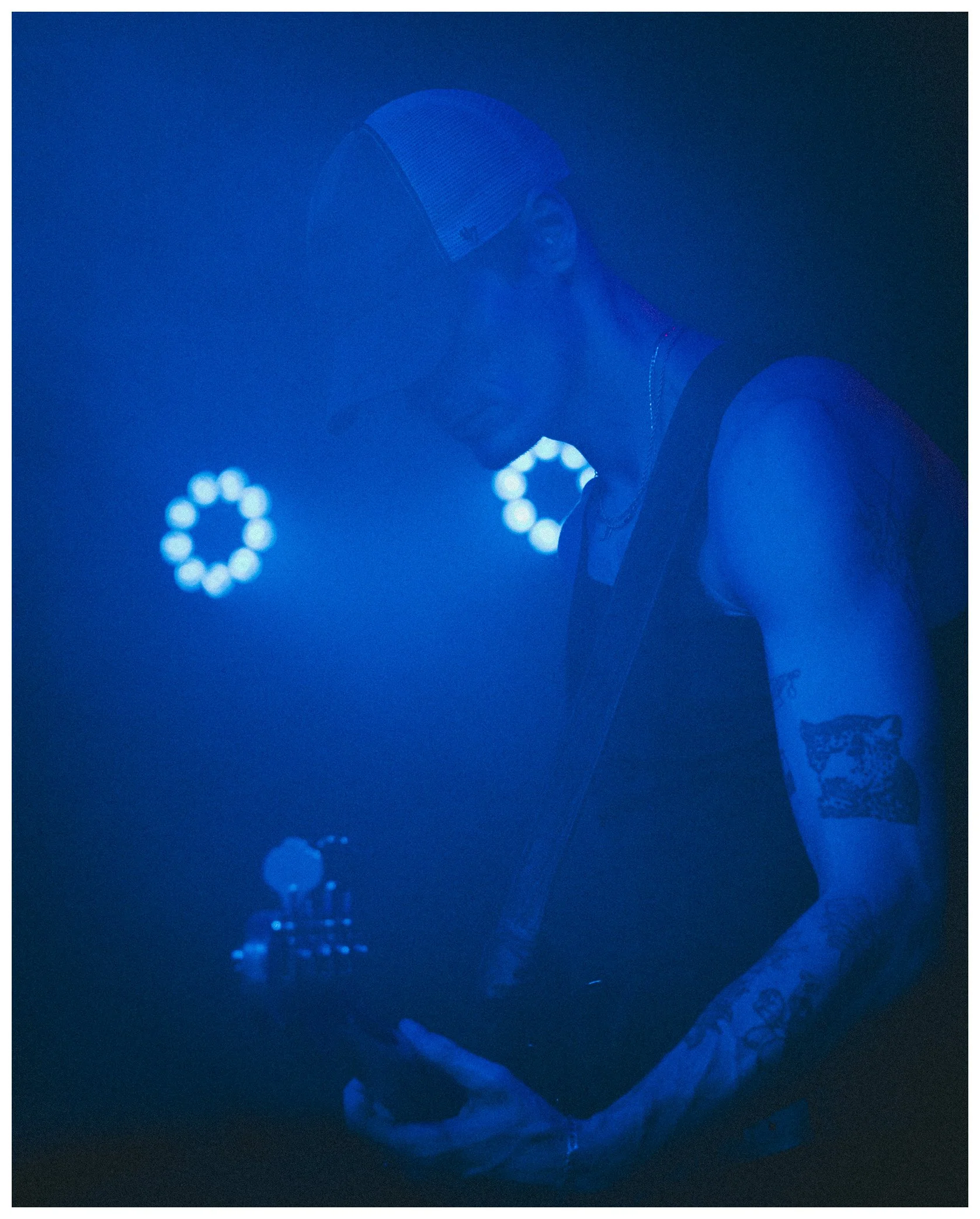 A person wearing a mesh cap and tank top stands under blue lighting, with a background of blurred circular light patterns.
