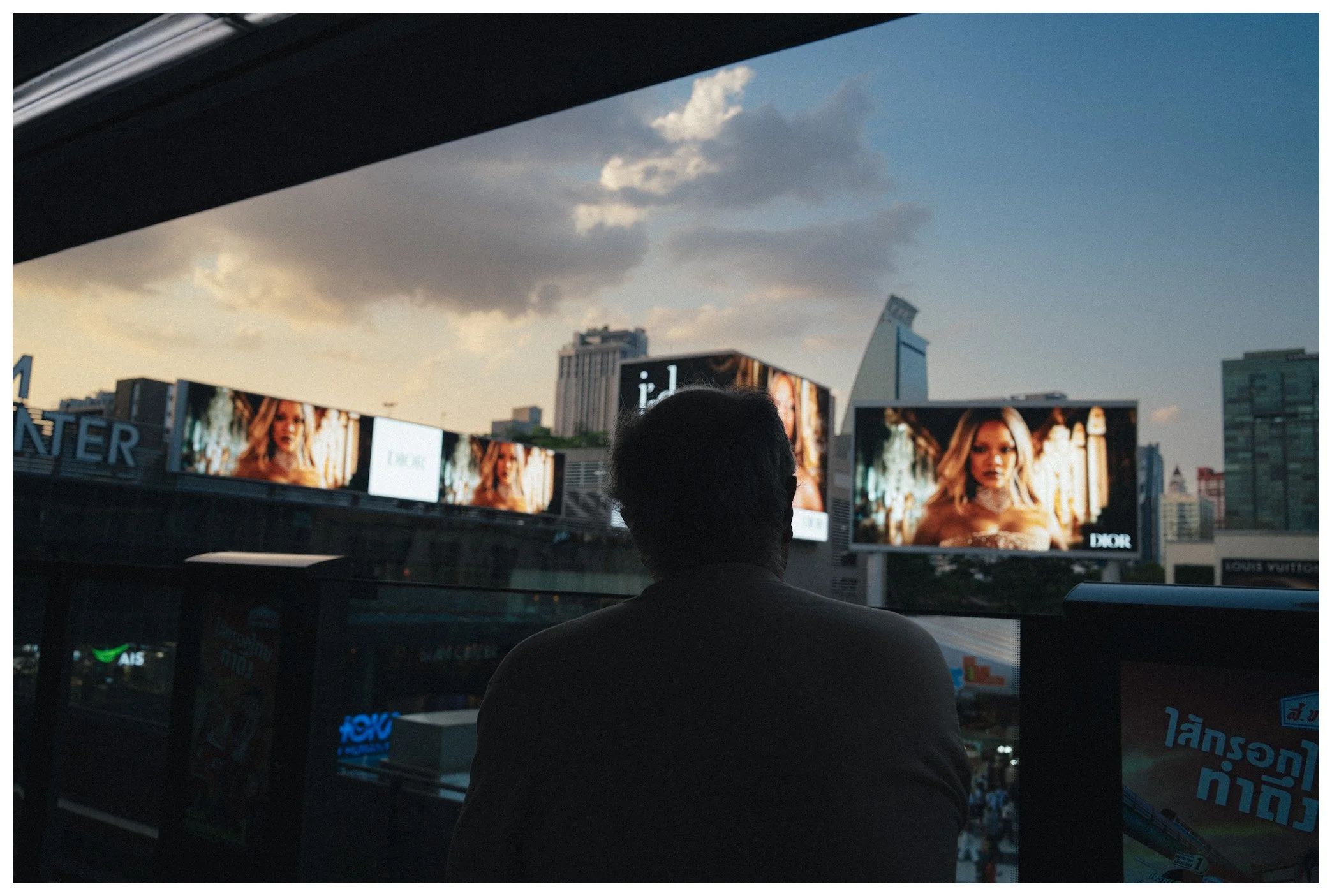 A person with short hair viewed from behind, standing in front of digital billboards displaying advertisements in a cityscape with tall buildings and a cloudy sky.
