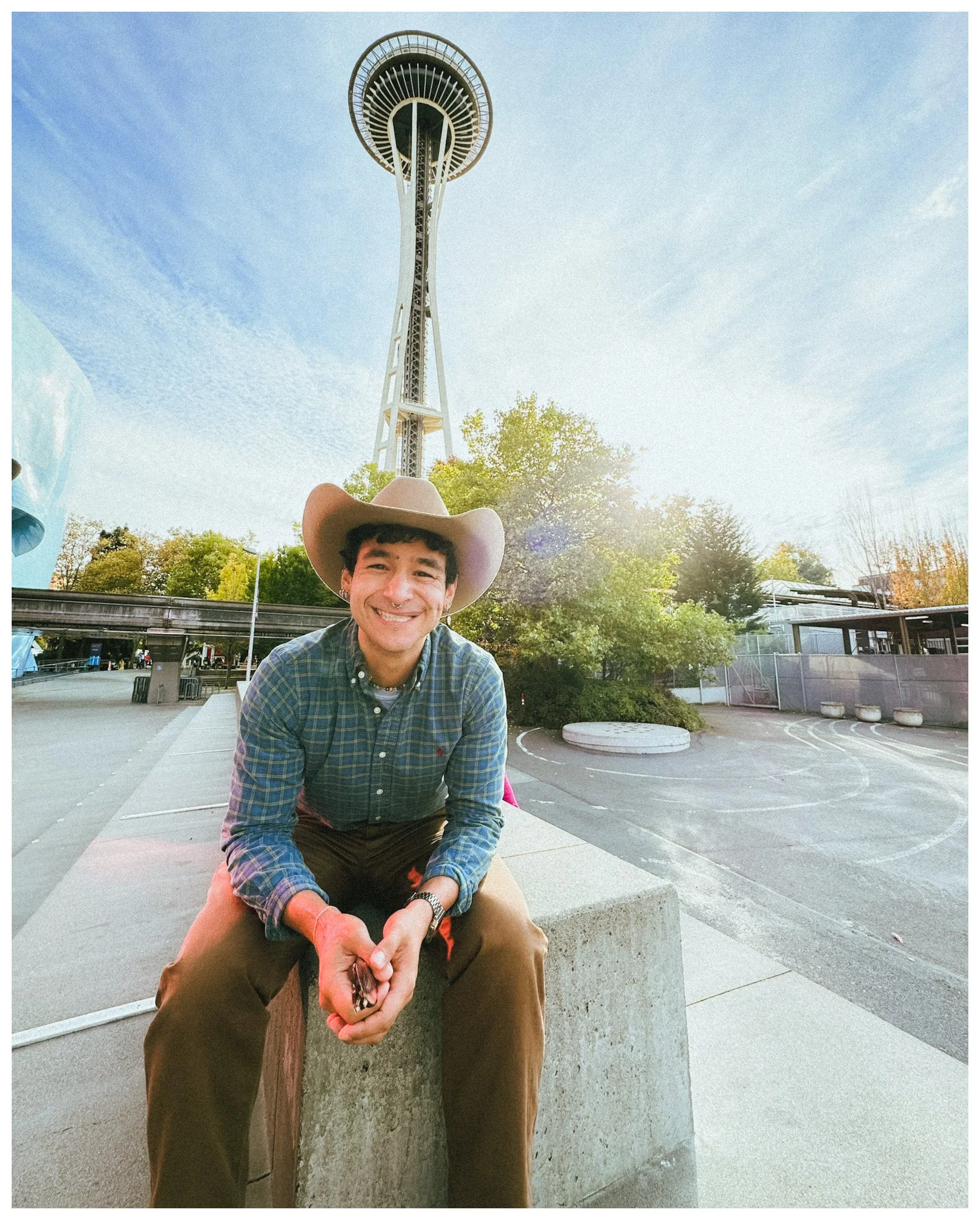 A smiling young man with dark curly hair and a hat crouching on a concrete ledge outdoors with the Space Needle in Seattle visible in the background.