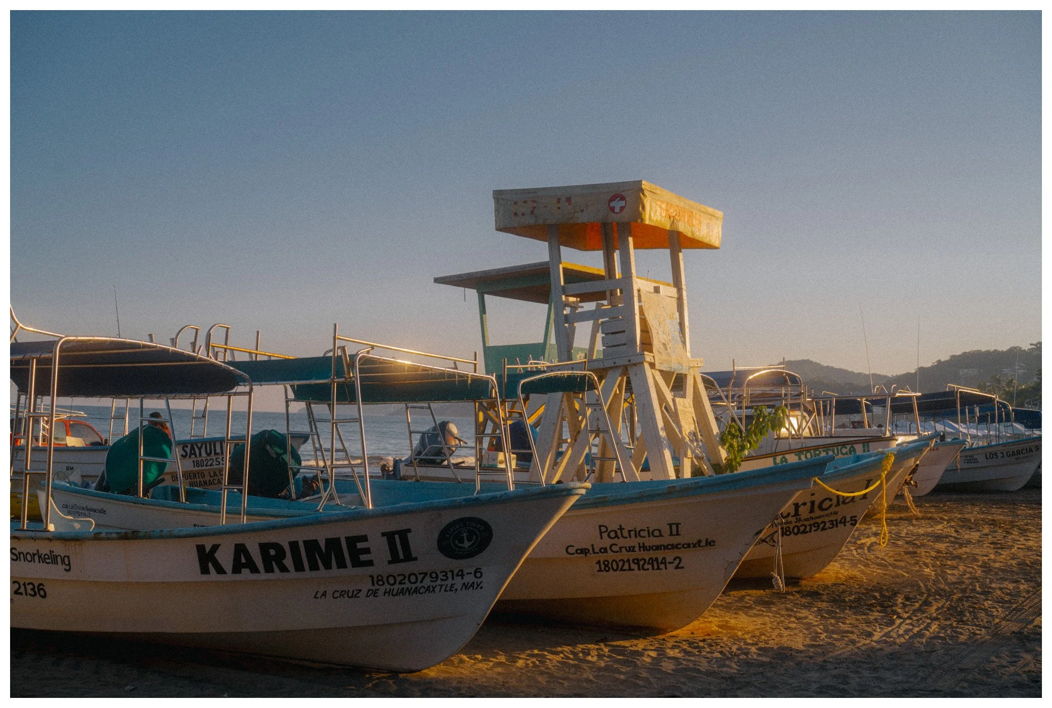 Boats docked on the shore with a lifeguard tower in the background, under clear blue sky during sunset.