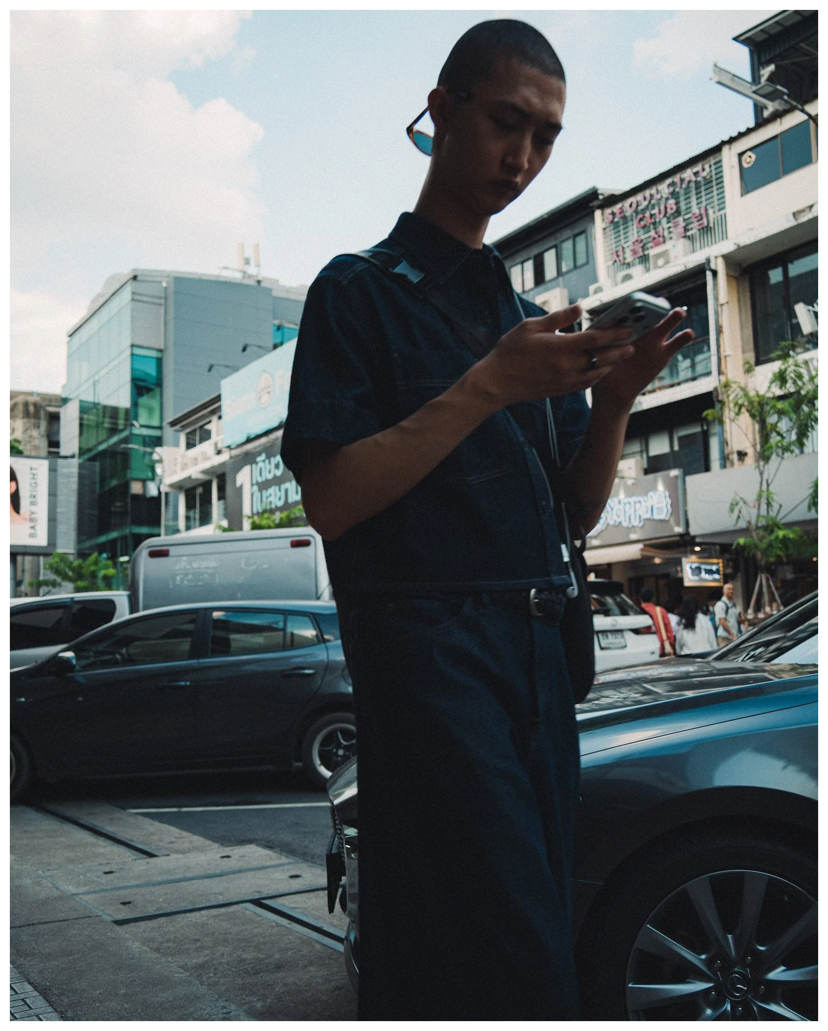 A man with short hair and glasses is standing on a city sidewalk looking at his phone. Behind him are several buildings with signs, cars parked and moving along the street, and pedestrians.