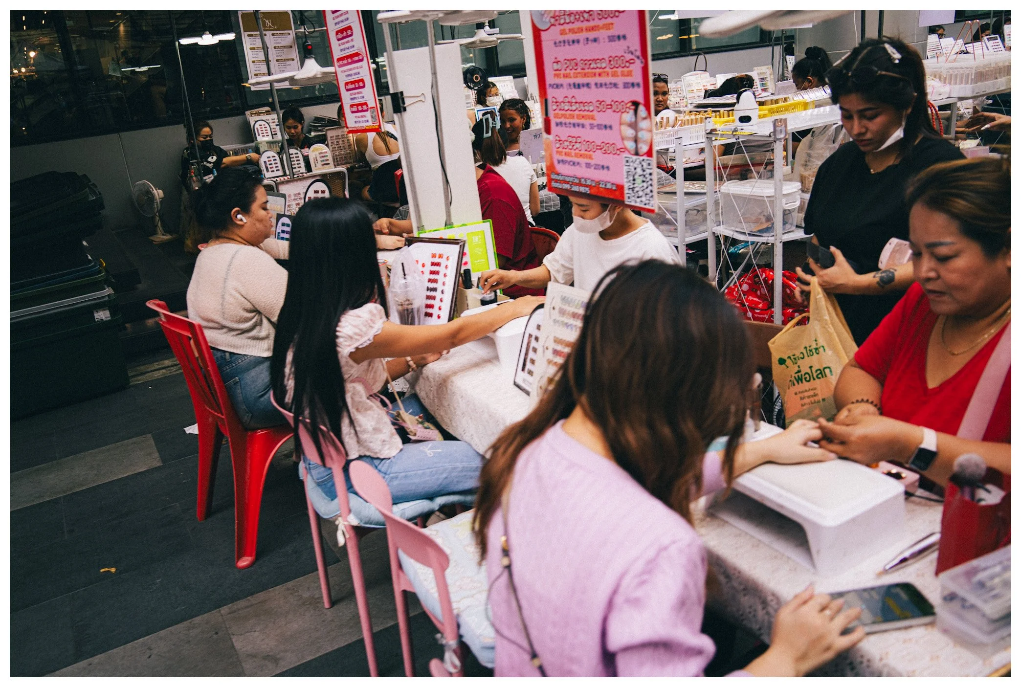 Group of people sitting at a long table inside a store or restaurant, with some wearing face masks and others using mobile phones, and a red promotional sign hanging from the ceiling.