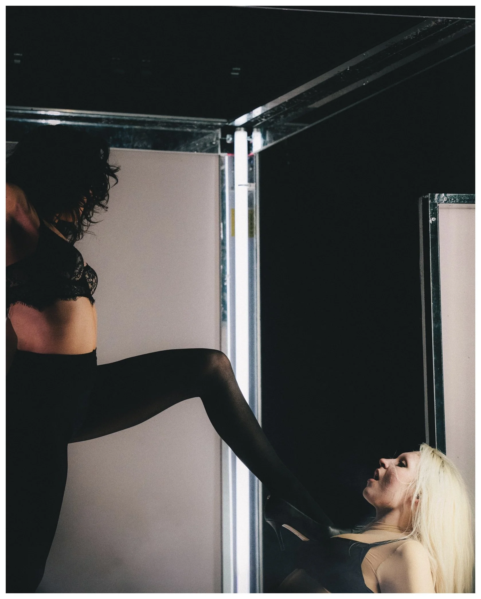 A woman with blonde hair stretching her leg against a wall, with her head tilted back, in a dance or fitness studio. A person with dark curly hair wearing a black lace top is partially visible on the left side of the image.