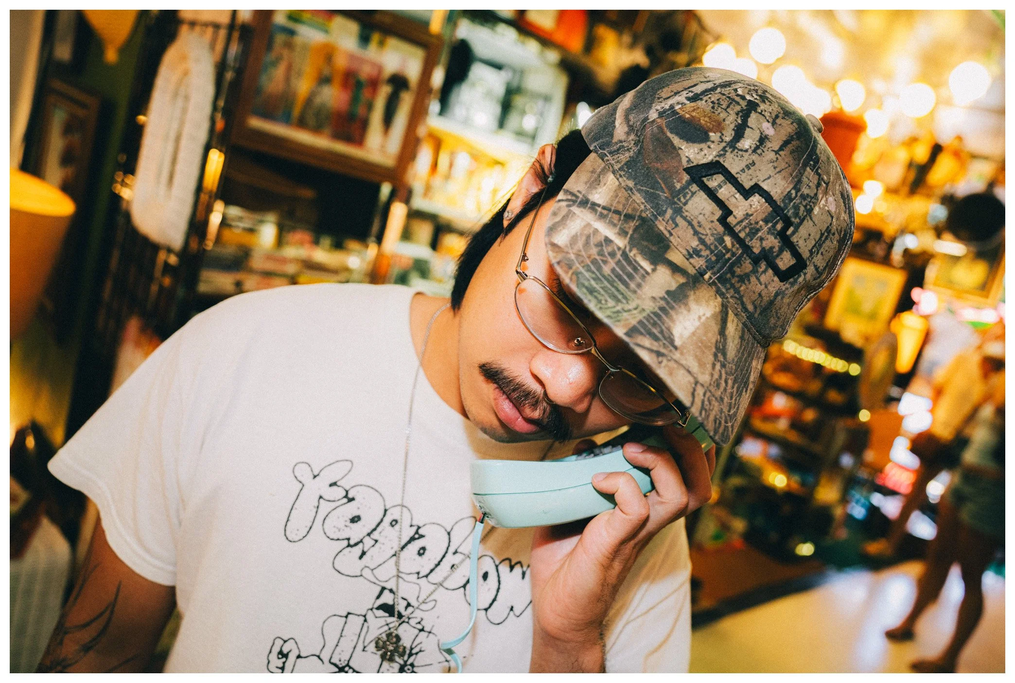 A man wearing a camouflage baseball cap and glasses is holding a light blue bowl with a white musical note design, and appears to be sniffing or tasting its contents inside a store filled with books and colorful decorations.