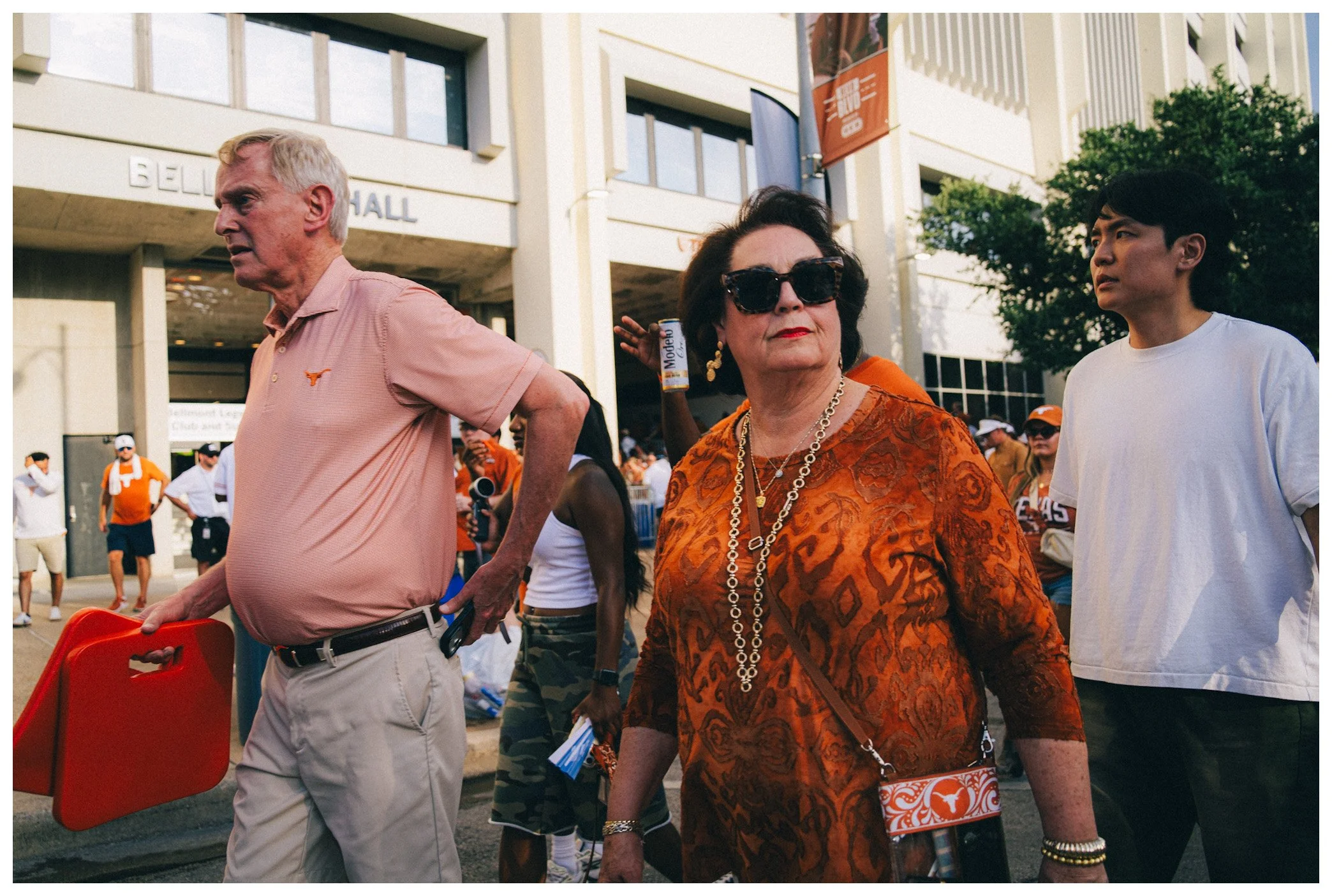 Group of diverse people walking outdoors in a city, with a building labeled 'BELL HALL' in the background, some carrying items and wearing casual clothing.