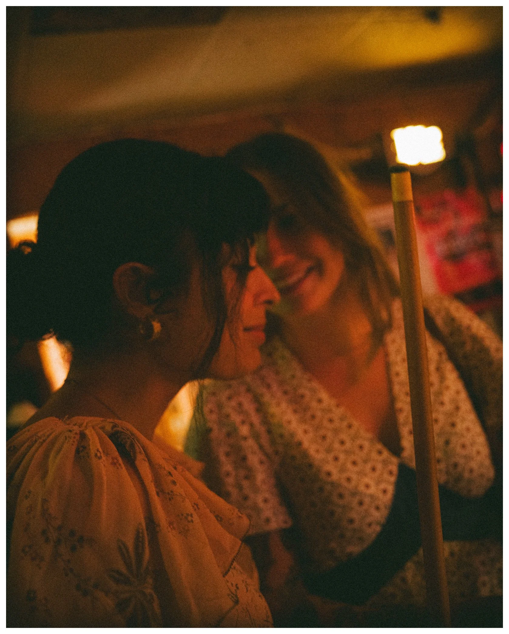 A man wearing a black cap and a woman with glasses smiling and leaning into each other in a dimly lit environment with warm lighting.