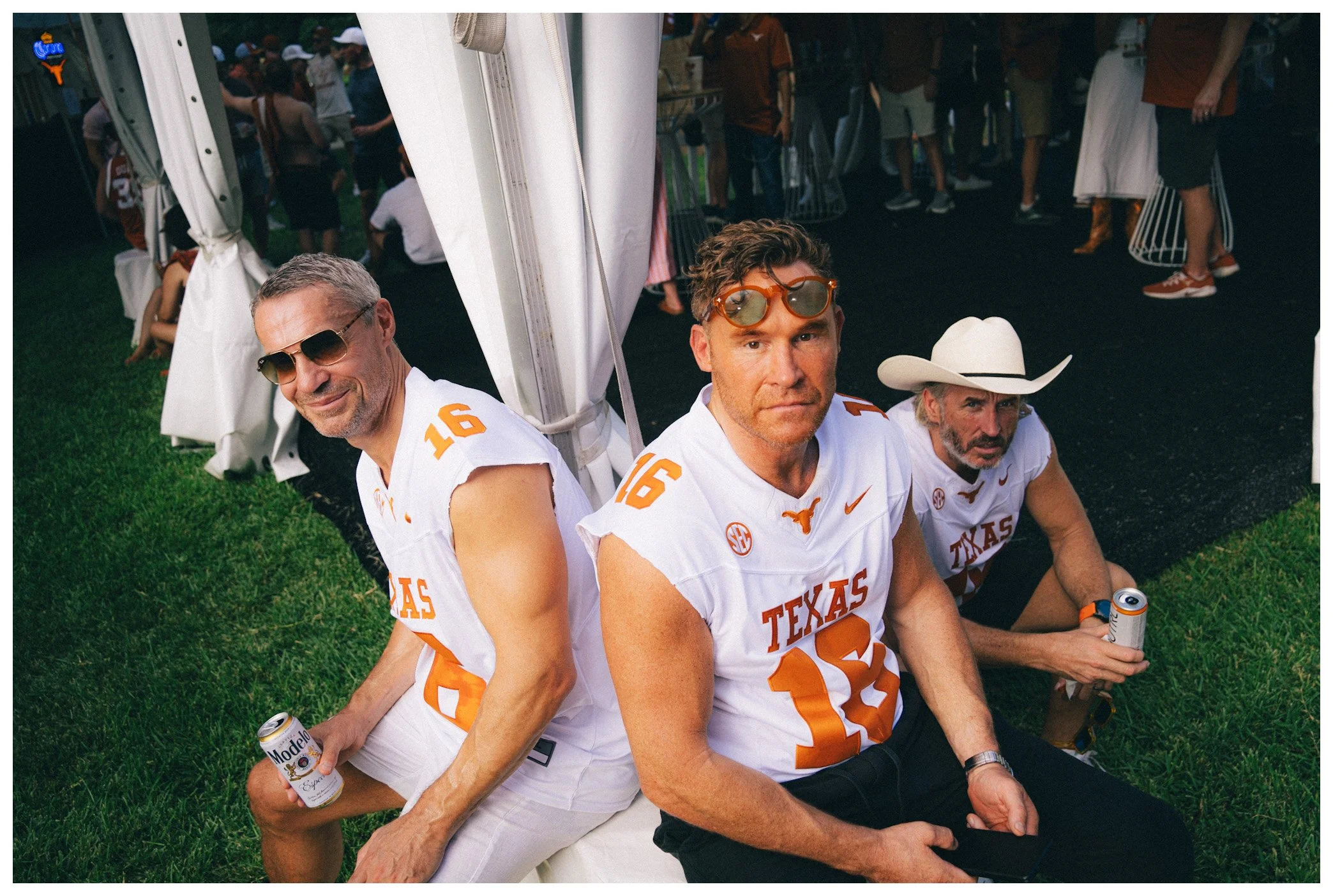 Three men sitting on the grass at an outdoor event, dressed in white and orange football jerseys, holding beer cans, with a crowd in the background.