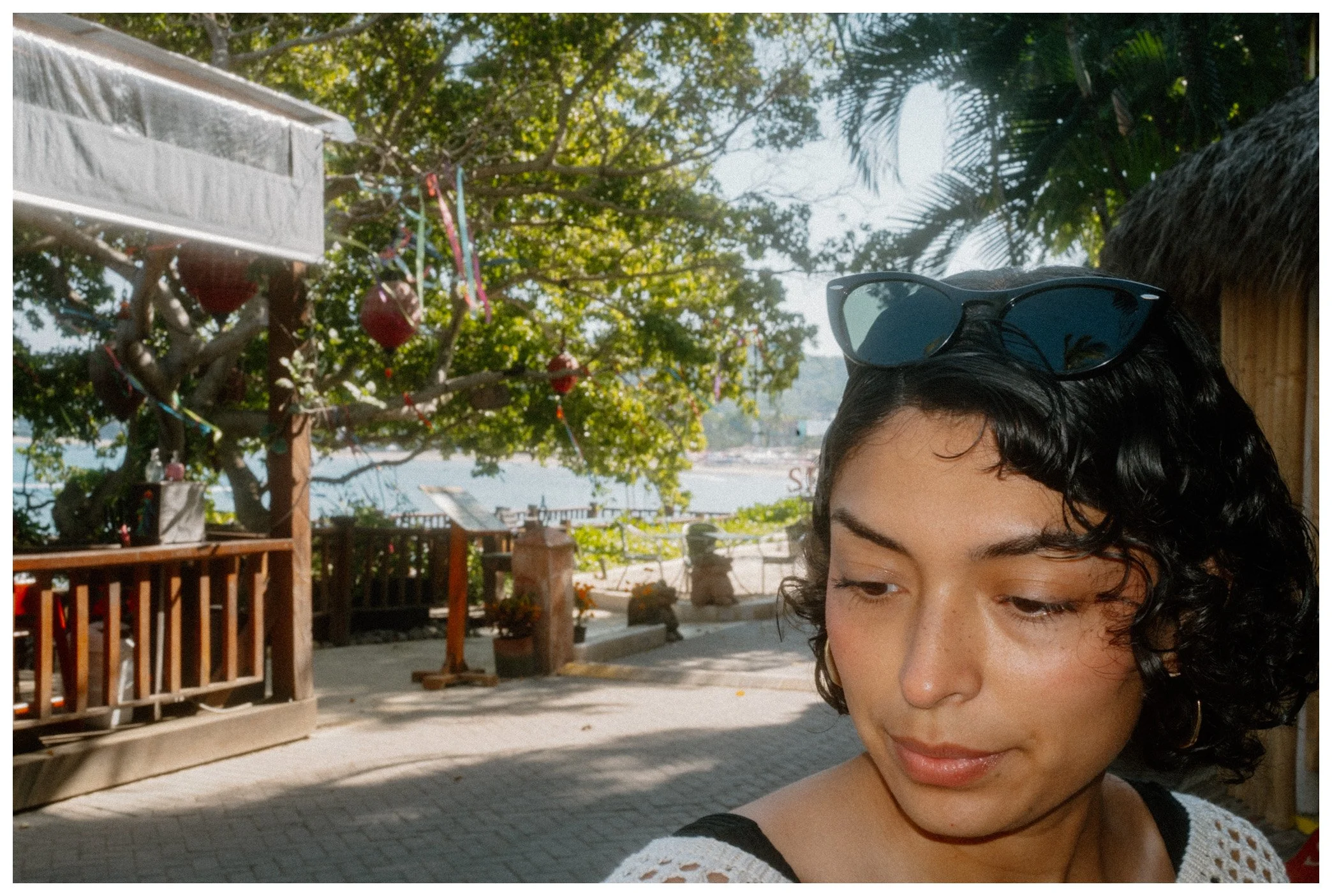 Close-up of a young woman with curly dark hair and sunglasses resting on her head, smiling gently outdoors with a background of trees, a beach, and a wooden deck.