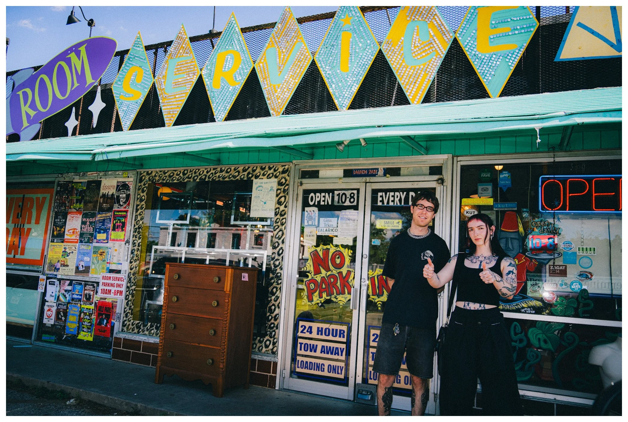 Diner exterior with colorful signs reading 'Room', 'Reservation', and 'Open daily 10-8', two young people standing in front, one with tattoos, one with glasses, smiling and giving thumbs up.