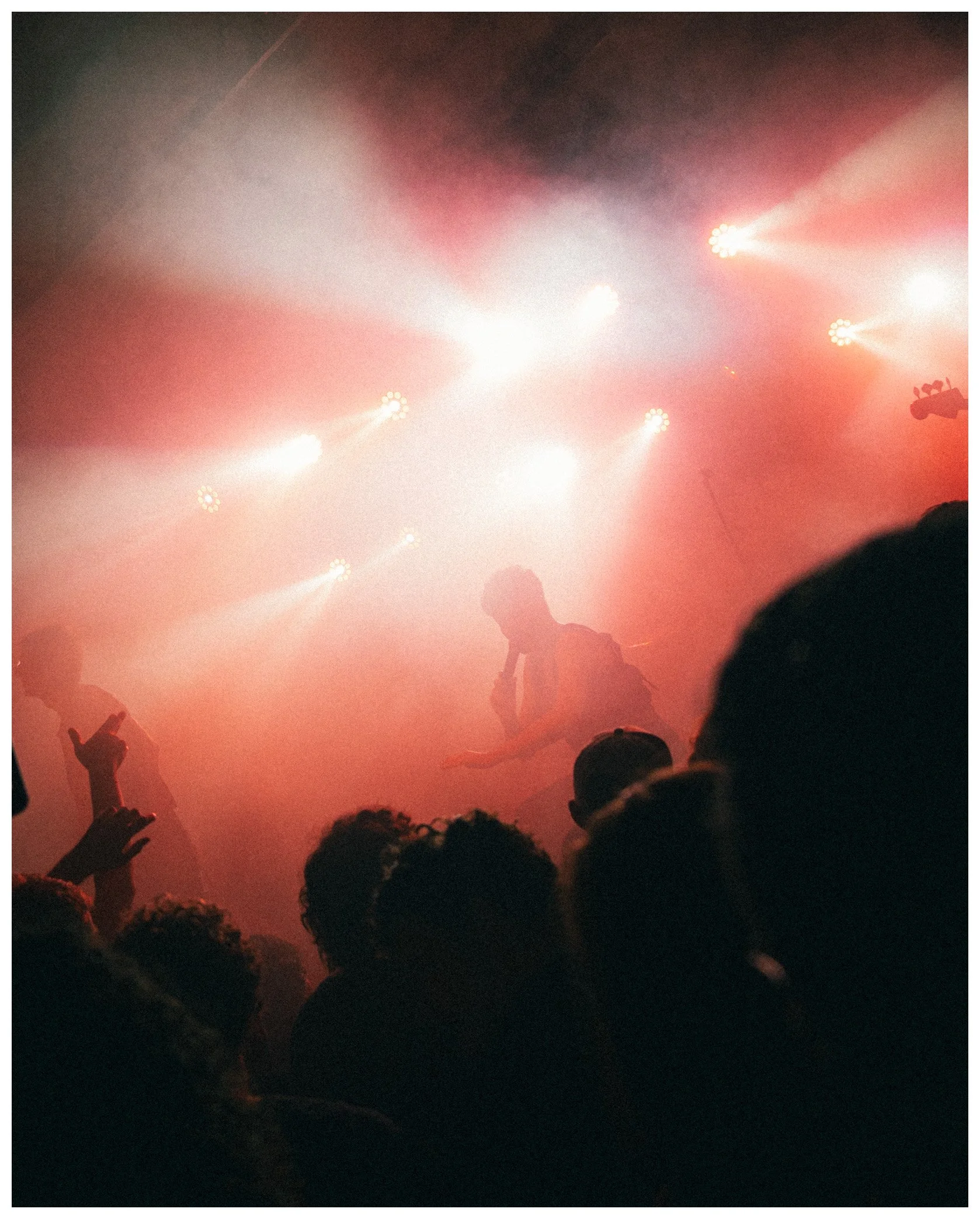 A crowd of people at a concert or event with bright stage lights and fog, with some individuals capturing the scene on their phones.